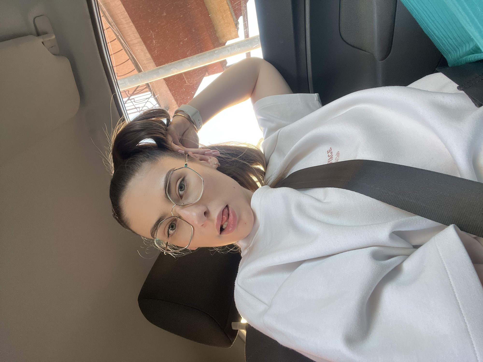 A person is positioned inside a vehicle, likely a car, as suggested by the presence of a seatbelt, a car seat, and a partial view of the car's interior including a sun visor. The individual has fair skin, dark hair pulled up, and is wearing eyeglasses. They are attired in a white sweatshirt or top. The person's arm is raised, possibly touching their hair or head. Outside the car window, a partial view of a building or structure is visible, appearing to be under construction or renovation, with scaffolding or metal beams in view. The precise location in the city of Unknown, Unknown is not identifiable from the provided image.