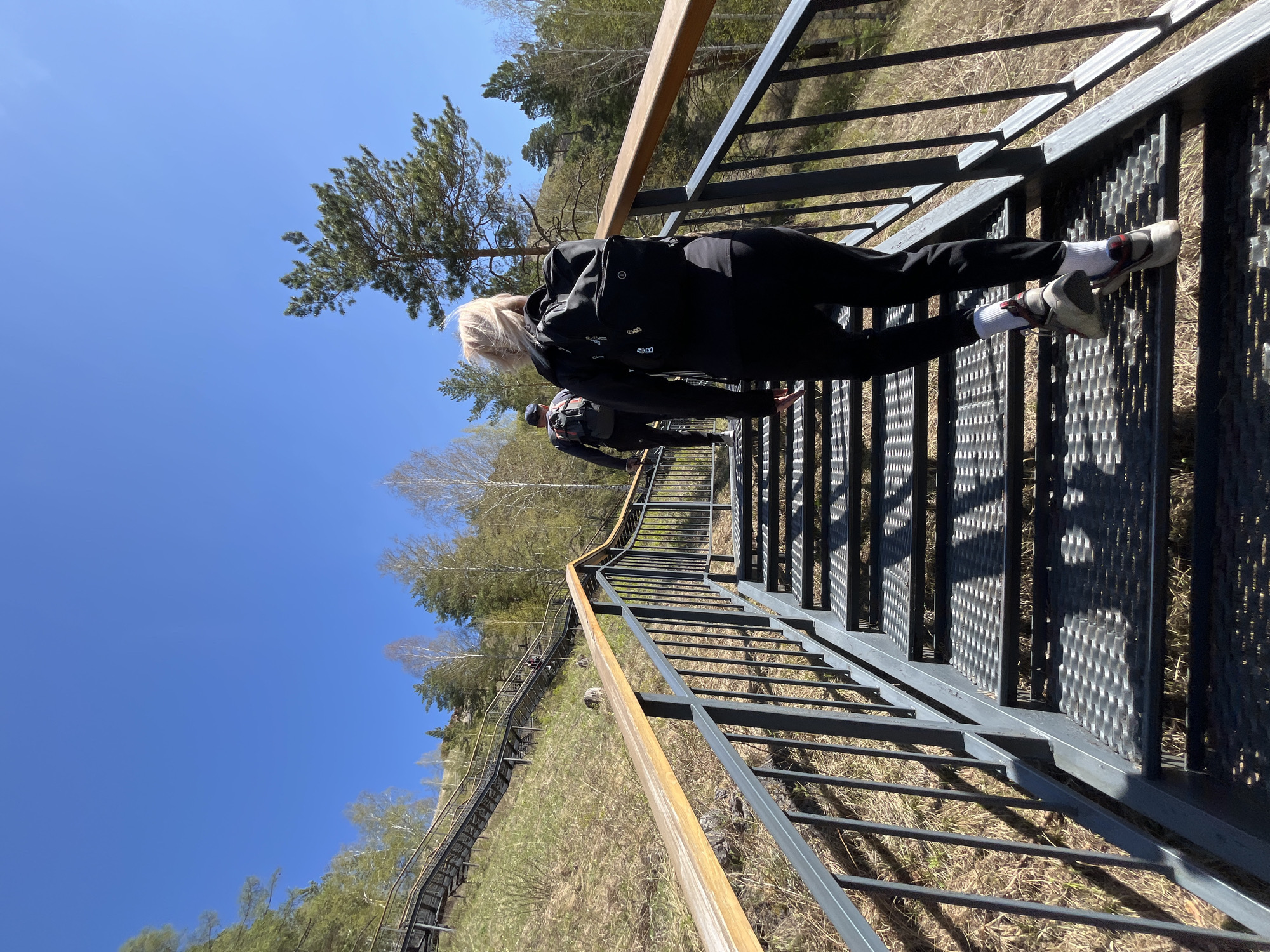 A medium, long shot captures people on a metallic staircase in Krasnoyarsk, Russia, under a clear blue sky. The woman in the foreground has blonde hair, wears a black tracksuit, white socks, and sneakers. She holds onto the railing and descends the stairs, her body slightly bent forward.
A man with a backpack follows behind her, also descending the stairs. The staircase itself is made of metal and has a wooden handrail. It winds down a grassy hill, with trees visible in the background. The shadows of the railing fall across the staircase.
