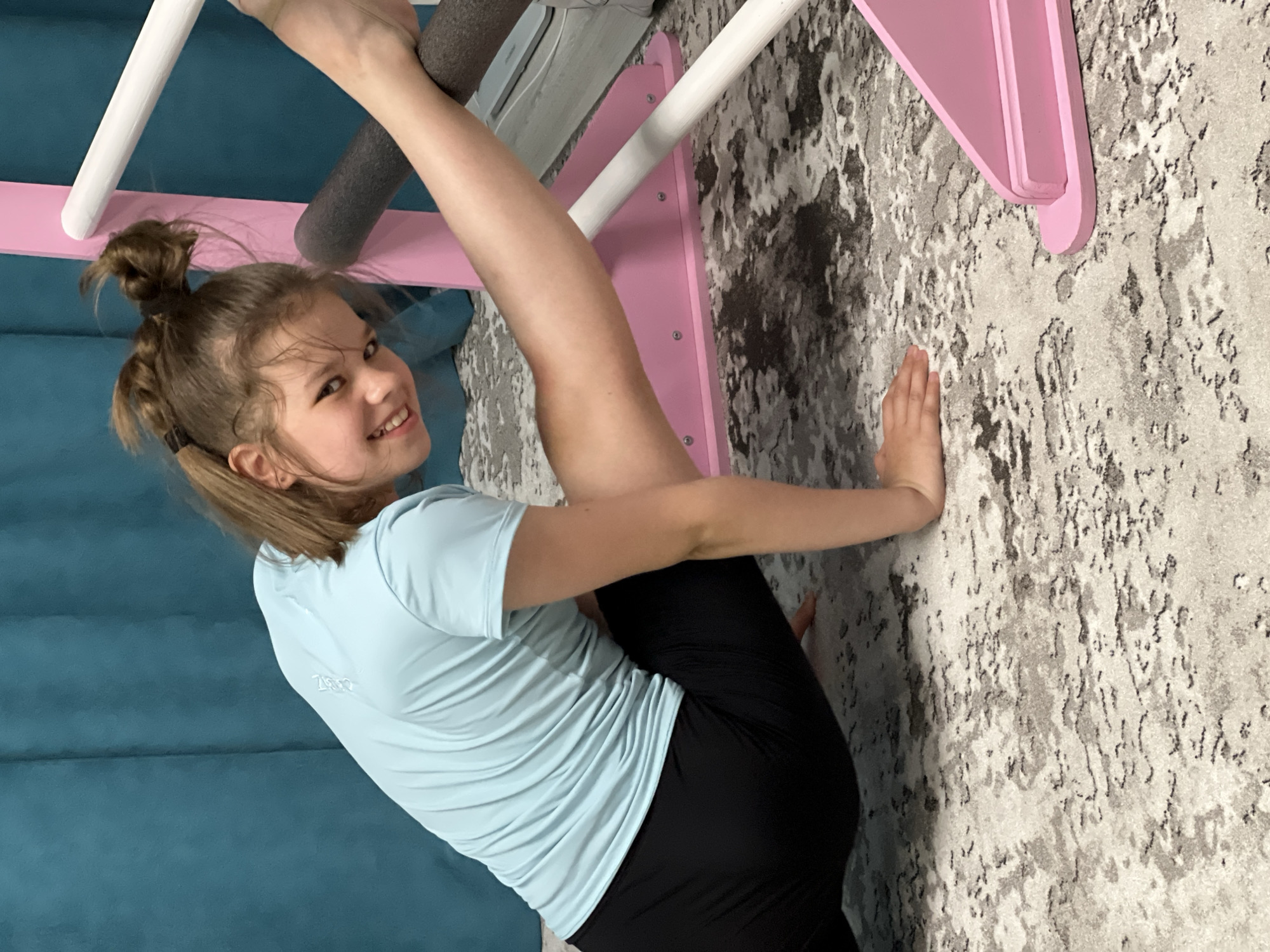 A young girl in a light blue shirt and black pants is performing a gymnastic pose on a Swedish ladder. She is holding onto the ladder with one hand and has one leg extended upward. Her other hand is placed on the wall for balance. The ladder is painted pink and white, and the wall has a textured gray and white pattern. The background includes a blue wall. The scene appears to be indoors, likely in a home. No specific location in the city of Chaikovsky, Russia is discernable.