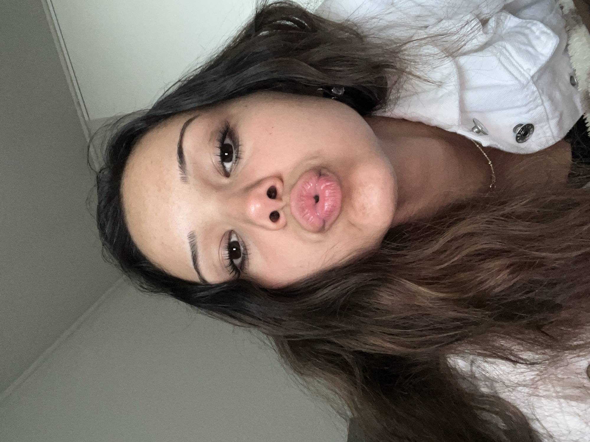 A close-up, head-on selfie photograph captures a young adult female indoors in the city of Unknown, Unknown. The subject has medium skin tone, dark brown eyes, and long, wavy dark brown hair with subtle lighter brown highlights, particularly visible on her left side (viewer's right). She is looking directly into the camera with her lips pursed into a "kiss" shape. Her eyes are open, and her eyelashes appear to have mascara applied. She wears a white denim-style jacket with visible silver snap buttons, a thin gold chain necklace, and small, clear or light-colored drop earrings on her right ear (viewer's left). Her hair frames her face and extends over her shoulders. The background is a plain, light gray wall, with a faint horizontal line visible on the ceiling in the upper left and a vertical white line, possibly a doorframe or architectural element, in the upper right. The scene is evenly lit.