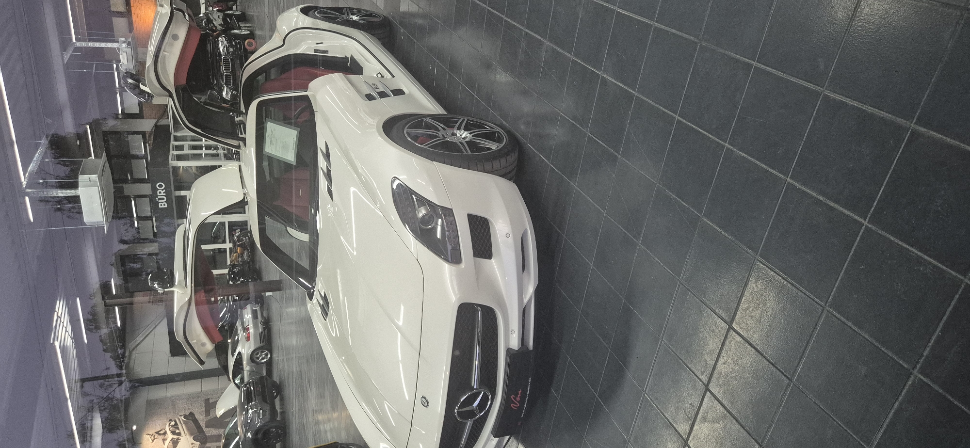 The image shows a white Mercedes-Benz SLS AMG with its gullwing doors open, parked indoors on a dark tiled floor. The car has red leather interior. In the background, other vehicles are visible, including a silver Porsche, a black SUV, and motorcycles. A sign that says "BÜRO" is visible in the background. The setting appears to be a car dealership or showroom. The car dealership is located in Mainz, Germany.