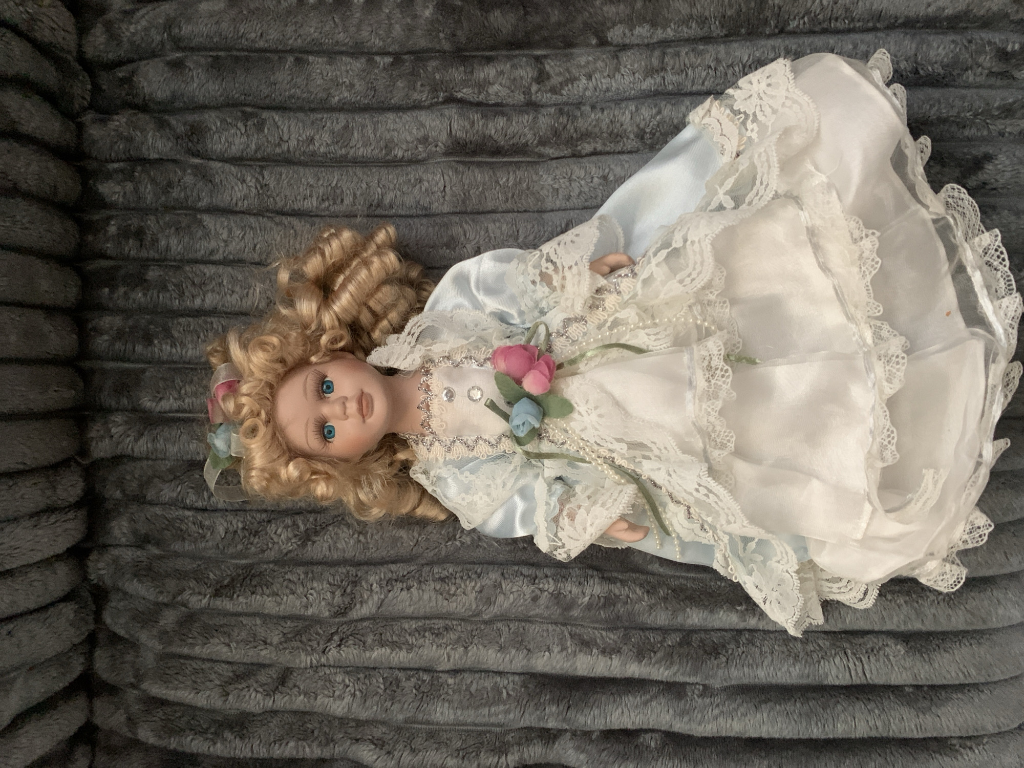 A porcelain doll with blonde curly hair and blue eyes is lying on a gray, textured blanket. The doll is wearing an elaborate dress with a white lace overlay and a light blue satin robe. There are small pink and blue fabric flowers attached to the front of the doll's dress, along with pearl beads. The doll's head is slightly tilted to the right. The background is entirely the gray blanket, which has a plush, ribbed texture. There is no indication of the city of Tashkent, Uzbekistan, or any urban environment in this image.