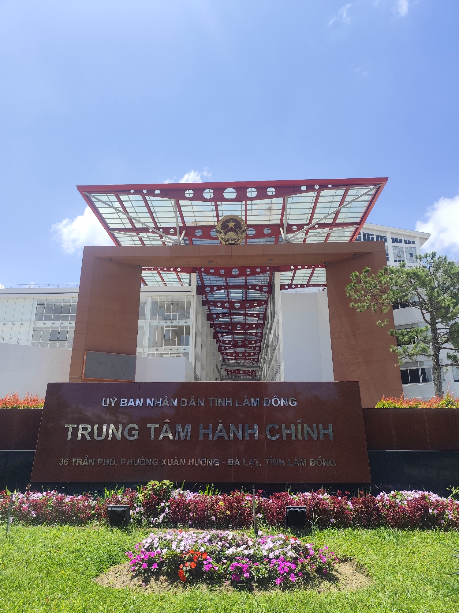The image shows the exterior of the Center of Administrative Services (TRUNG TÂM HÀNH CHÍNH) in Da Lat, Vietnam, located at 36 Tran Phu Street, Xuan Huong Ward, Da Lat City, Lam Dong Province. The building features a modern architectural design with a prominent red metal frame supporting a glass roof over the entrance. The facade is clad in warm brown panels. In front of the building, a large, dark-colored sign displays the name of the center and its address in Vietnamese, with reflective silver lettering. The foreground is adorned with meticulously maintained flower beds containing a variety of colorful flowers in shades of pink, purple, red, and white, interspersed with lush green grass. A single black outdoor light fixture is visible amongst the flowers. The sky above is a clear blue with a few wispy white clouds, suggesting a bright, sunny day. There are no people visible in the image. The overall impression is one of an official, well-kept public building.