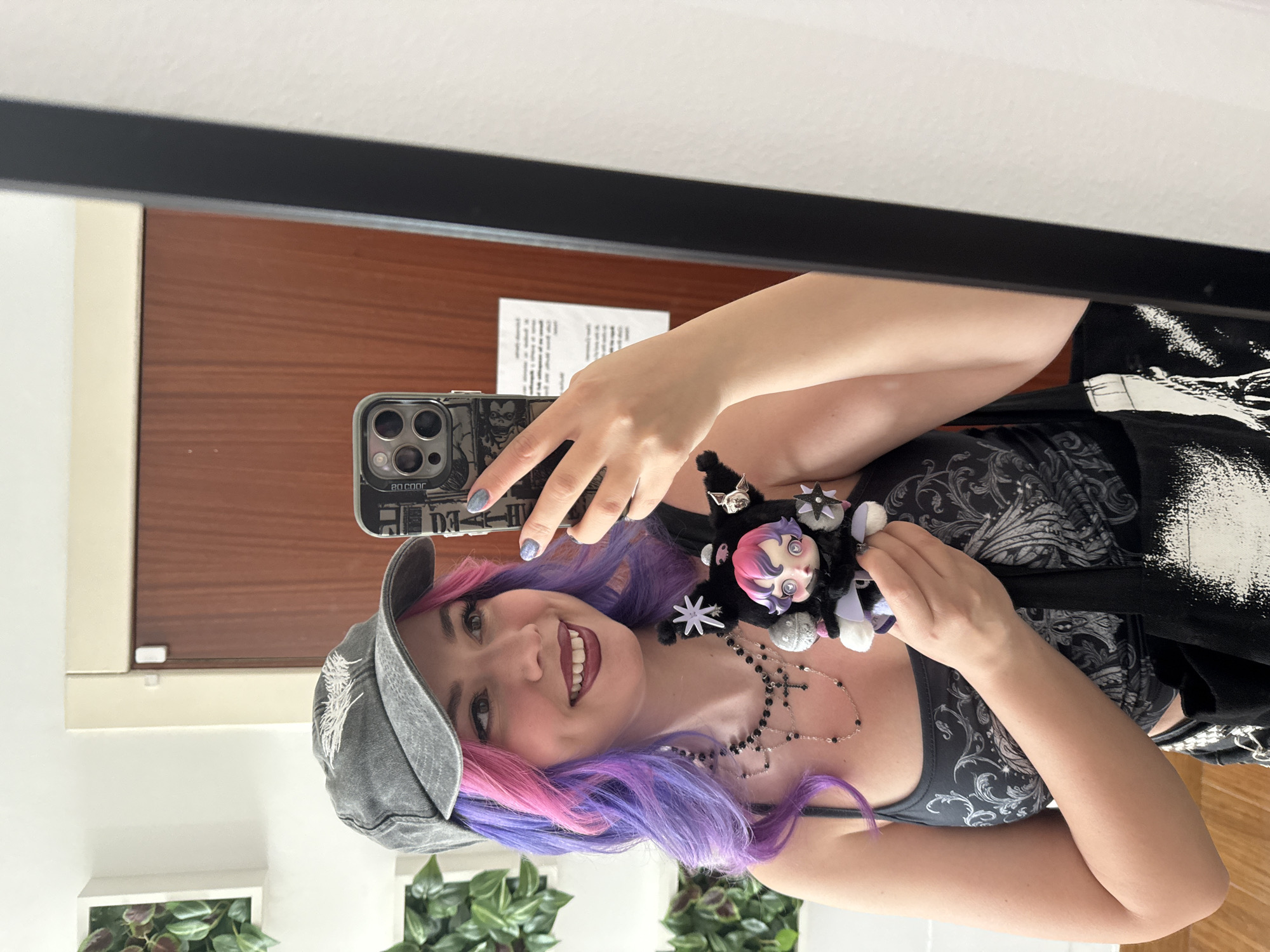 A woman stands in front of a mirror, holding her phone to take a selfie. She wears a gray baseball cap, a black tank top with a pattern, and necklaces. Her hair is dyed pink and purple. In her other hand, she holds a black doll with a pink-haired head. The reflection in the mirror shows a light-colored wall, a dark wood-paneled door, and a framed plant. A paper hangs on the wall near the door. The environment suggests an indoor setting, possibly a room in a building. There is no indication of the location being in Madrid, Spain.