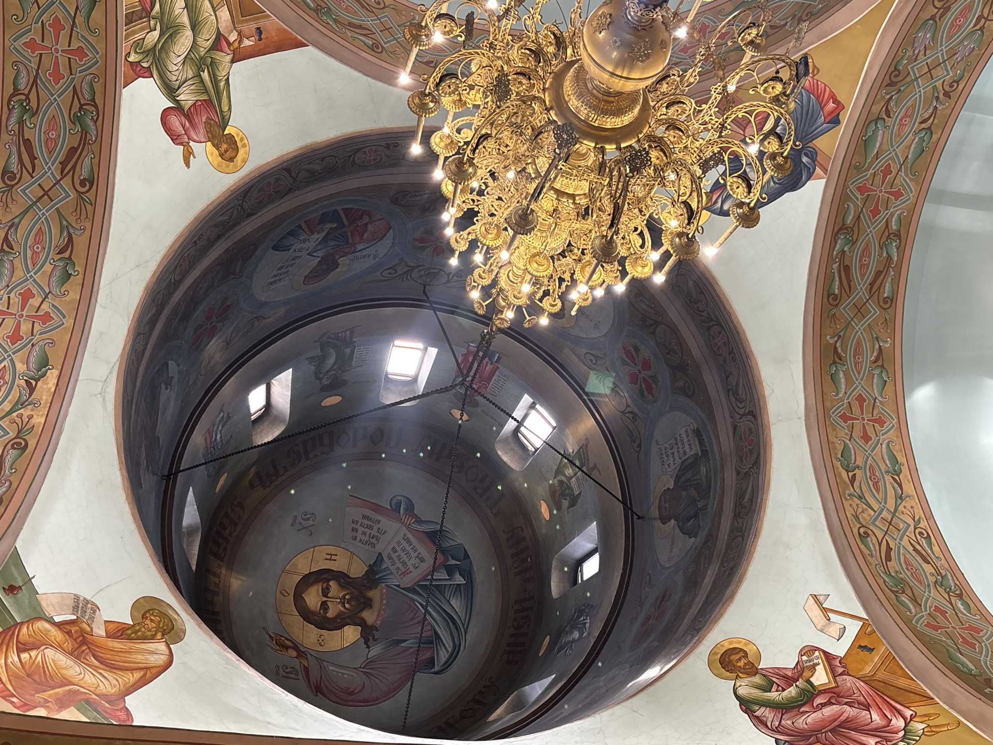 The image captures an upward view inside a church in the city of Chaikovsky, Russia, showcasing the interior of a dome. The dome's surface is adorned with religious iconography, including images of Jesus Christ and other biblical figures. Some of these figures are surrounded by halos.
Hanging centrally beneath the dome is an ornate, multi-tiered chandelier, emitting light. The dome features several small windows. The surrounding ceiling is painted with decorative patterns.