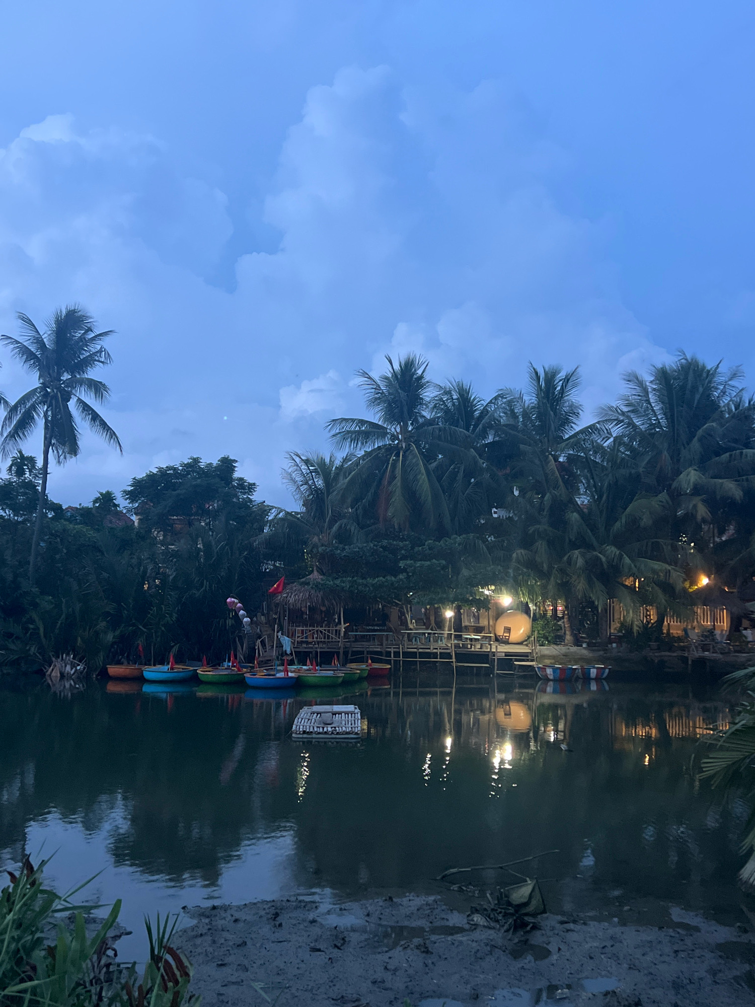The image depicts a serene canal scene in Hoi An, Vietnam, likely during twilight. The water's surface reflects the soft glow of lights from buildings nestled among lush palm trees lining the canal. A row of small, brightly colored boats is moored along the bank, their reflections shimmering in the water. A thatched-roof structure with wooden railings and tables suggests a waterside establishment. The sky above is a deep blue, with scattered clouds adding texture. The foreground shows a muddy bank with some vegetation. The overall atmosphere is tranquil and picturesque, characteristic of Hoi An's charming waterways.