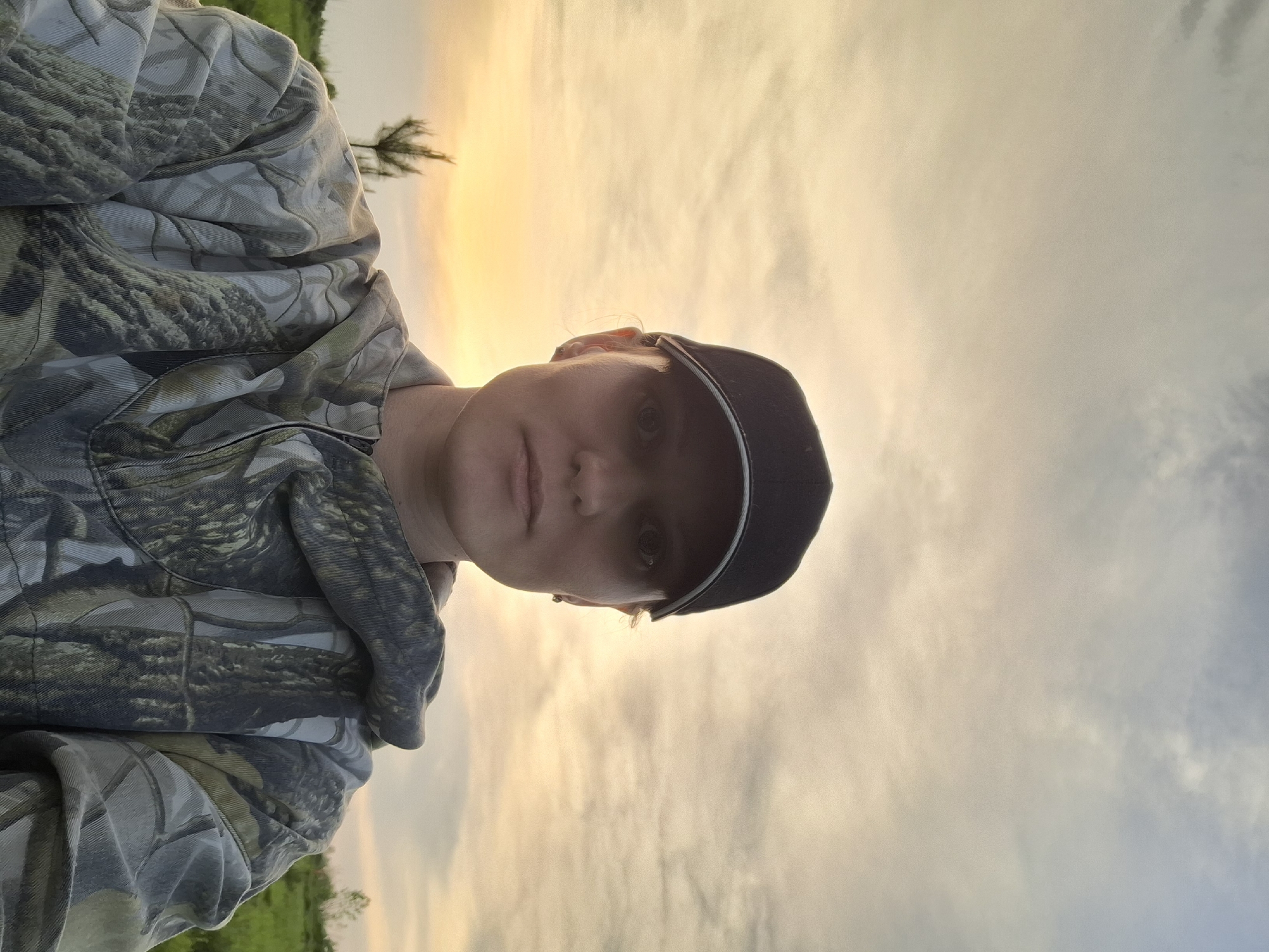 A person wearing a camouflage hooded sweatshirt and a dark baseball cap is the primary subject. The background features a sky with streaks of orange and white, suggesting either sunrise or sunset. Below the sky, a strip of green vegetation is visible. The photo appears to be taken outdoors in a natural setting; there is no indication of the location being in the city of Шешурга, Russia.