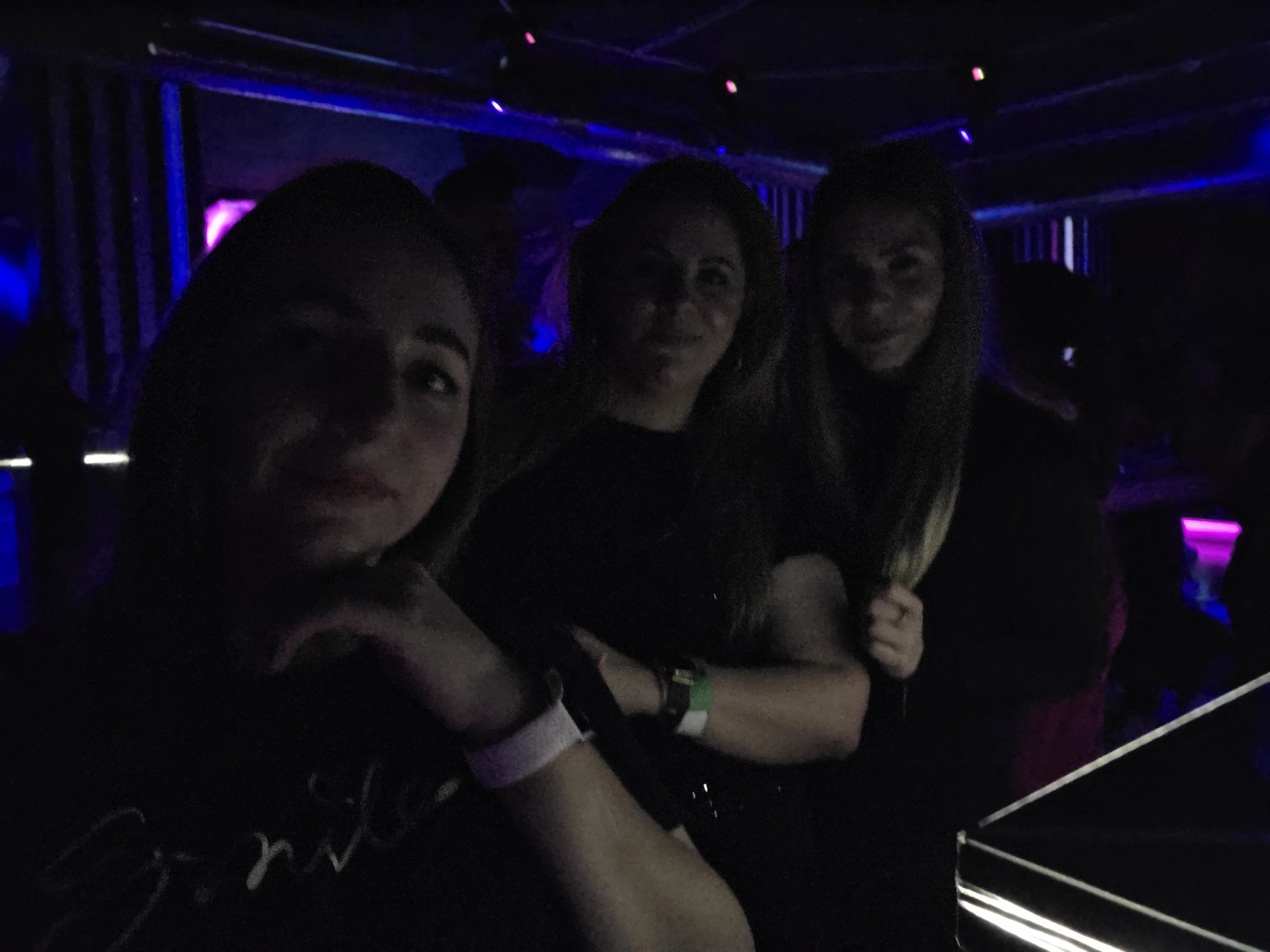 Three young women pose for a selfie in a dimly lit environment, potentially a nightclub, evidenced by the colorful lighting and indistinct figures in the background. The woman in the foreground is on the left, with her left hand raised near her face, and her wrist has a light-colored wristband. To her right are two other women, both also facing the camera. All three women are wearing dark clothing. In the background, there are structural elements that appear to be metal bars or supports, and several sources of light are present, including blue and pink lights. A shiny, reflective surface is present in the lower right corner.