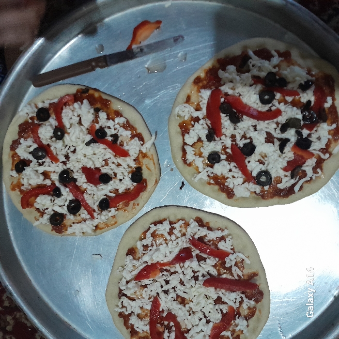 The image depicts three uncooked mini pizzas arranged on a large, round, silver-colored metal tray, set within a domestic environment in Khan Yunis, Palestinian Territory. Each pizza features a light-colored dough base topped with a layer of red tomato sauce, a substantial amount of shredded white cheese, scattered black olive slices, and several strips of red bell pepper. To the upper right on the tray, a knife with a dark brown handle and a silver blade is present, with a small piece of red bell pepper adhering to its tip, indicating recent use for ingredient preparation. The metallic tray shows minor wear and surface reflections. A dark, patterned rug or textile is partially visible in the upper left corner of the background. A "Galaxy A14" watermark is present in the lower left corner of the image.