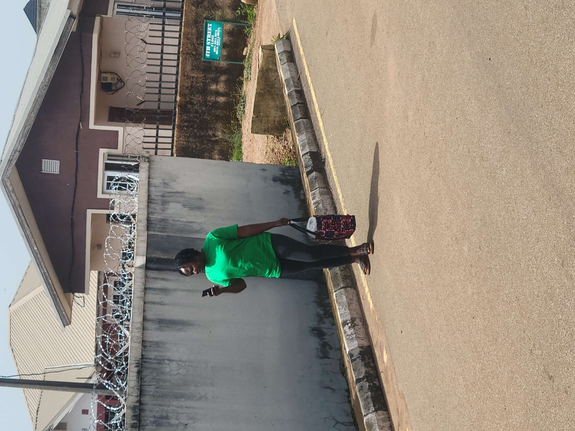 A woman stands on a paved road in front of a concrete wall topped with barbed wire fencing. She is wearing a bright green t-shirt, black leggings, and dark sandals. In her right hand, she holds a mobile phone, and in her left hand, she carries a patterned tote bag. The road has a yellow line painted along the curb, which is made of concrete blocks. Behind the wall, a multi-story building with a brown and beige facade is visible, as well as a beige-colored roof. A green sign that reads "4TH STREET CLOSE" is mounted on the wall to the right of the woman. The sky is clear and bright, suggesting a sunny day. This scene is located on 4th Street Close in Jos, Nigeria.