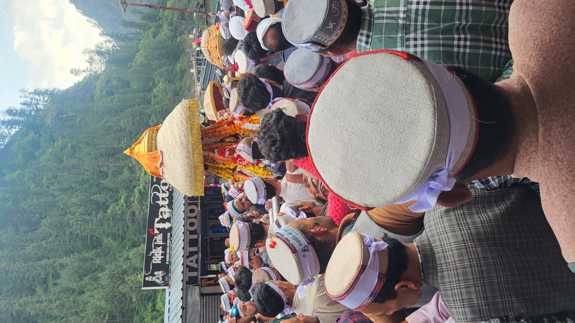 The image depicts a vibrant cultural or religious procession unfolding on a street in a mountainous region, likely Manikaran, India. A large crowd of men fills the scene, mostly viewed from behind or above, indicating their participation or observation of the event. Many individuals are wearing traditional Himachali caps, characterized by their light colors, some with red or patterned bands, and others featuring white bands with Devanagari inscriptions.
At the heart of the procession is a grand, palanquin-like structure, possibly carrying a deity. It is elaborately adorned with abundant garlands of yellow and orange marigolds and other colorful flowers, topped with a golden/yellow canopy, and rests on a prominent, rounded, white/cream base with gold fringing. Another similar but smaller structure is visible further back in the crowd. Many attendees are holding up mobile phones, suggesting they are recording or capturing photographs of the event.
The scene takes place outdoors during the daytime under a bright, partly cloudy sky. The background is dominated by towering, lush green forested mountains, typical of the Himalayan landscape. Buildings line one side of the street, including one that hosts a tattoo parlor. A prominent sign for "Rink Ink Tattoo" is visible on this building, featuring the text in English and its Devanagari equivalent "रंक इंक टैटू", along with phone numbers (99688 81655, 97360 01133). The word "TATTOO" is also prominently displayed. Some of the Himachali caps feature legible Devanagari script, such as "नारायण श्री देवि" (Narayan Shri Devi), and an "ॐ" (Om) symbol is faintly visible on another cap. The overall impression is a bustling community gathering for a significant cultural or religious occasion in a picturesque mountain town.