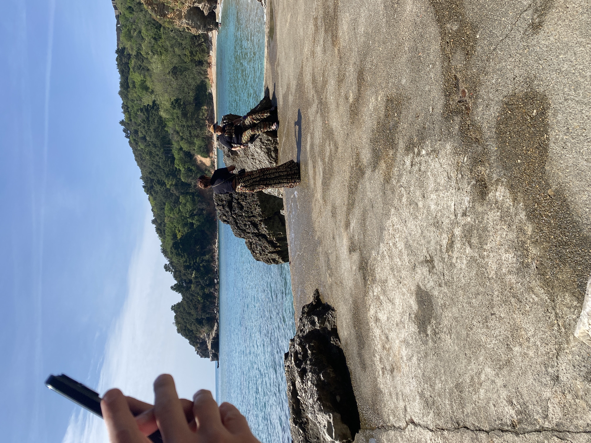 The scene is captured outdoors with a view of the sea and a rocky coastline, likely in the vicinity of Budva, Montenegro. Two individuals are positioned along the rocky shore. The foreground features a concrete surface, slightly cracked. The perspective suggests someone is taking a picture. The backdrop includes clear blue skies with scattered clouds, a lush green hillside along the coast, and the sea.