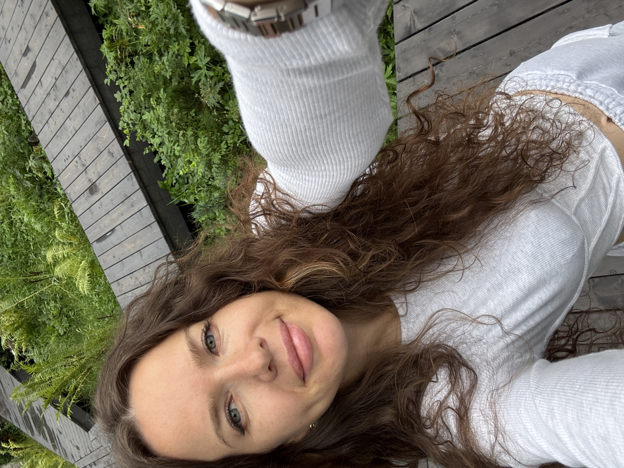 A medium shot captures a young woman with long, wavy brown hair, light blue eyes, and a light complexion. She is wearing a white long-sleeved shirt and is looking directly at the camera, smiling softly. Her left arm is extended, holding a smartphone for a selfie. The background consists of a wooden walkway and lush green vegetation, suggesting an outdoor environment. The scene is set on the outskirts of Nizhnekayancha, Russia, where wooden pathways are often integrated into natural landscapes for pedestrian access.