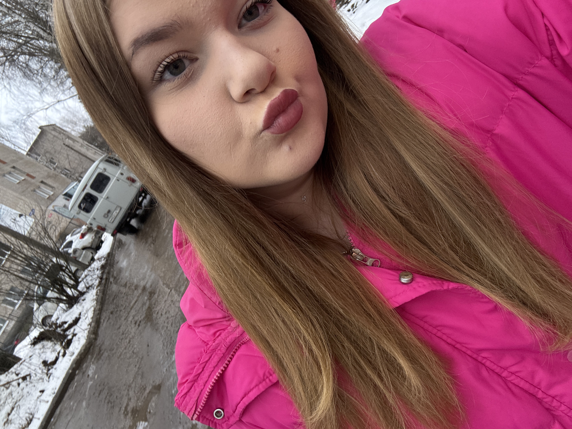 A young woman with long blonde hair and light skin is taking a selfie. She is wearing a bright pink jacket and making a kissing face. The background shows a street scene in what appears to be Chaikovsky, Russia. Patches of snow are visible on the ground, and a white van and other cars are parked on the street. A grey building is in the background.