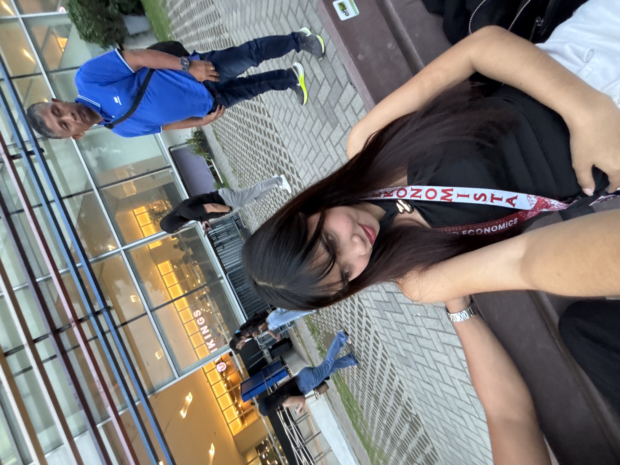 The image shows a group of people outside a building identified as "KIINGS" at Ateneo de Davao University in Davao City, Philippines. A man in a blue shirt and jeans stands near the building, while other people are seated nearby. A woman in a black top is visible in the foreground wearing a lanyard with the word "ECONOMISTA." The environment appears to be a paved outdoor area.