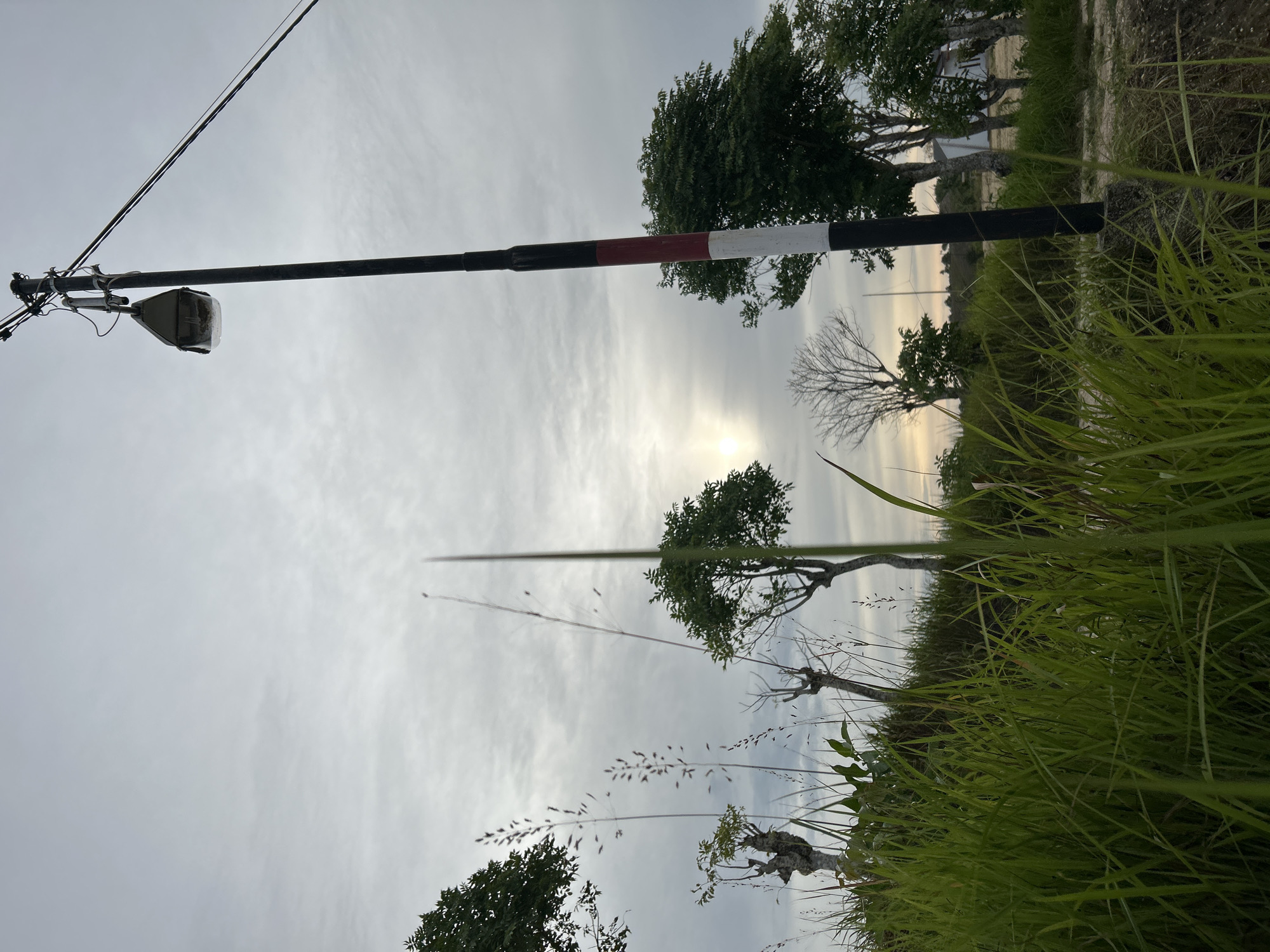 The image, possibly taken in Lhokseumawe, Indonesia, shows an overcast outdoor scene with a focus on a black and red-white striped boom barrier extending from a utility pole on the left towards the center. A street light hangs from the pole. The foreground is filled with tall green grass and some low-lying vegetation. In the background, there are trees and what appear to be buildings or structures. The sky is cloudy with the sun faintly visible behind the clouds. The perspective is from a low angle.