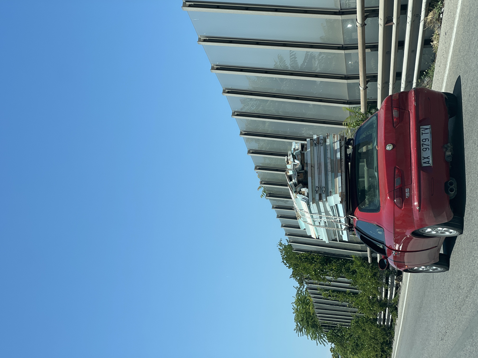 A red Alfa Romeo 155 is driving on a road next to a sound barrier. The car is loaded with a stack of large, flat objects on its roof rack. The objects appear to be made of a light blue insulating material with a metallic trim. The car has a license plate that reads "AX 979 TV". The sound barrier is made of vertical metal panels with a frosted glass-like insert. Trees and greenery are visible behind the sound barrier. The sky is clear and blue. The scene appears to be in a highway or a large road, possibly on the outskirts of Trani, Italy.