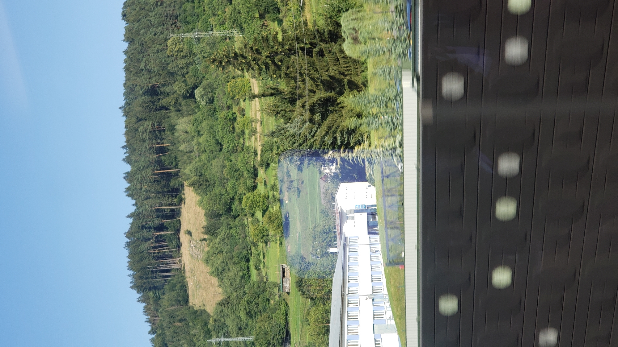 The image depicts a sunny day with a clear blue sky above a densely wooded hillside. The hillside is covered with a mix of evergreen and deciduous trees, with some patches of dry grass visible on the slope. In the mid-ground, there is a long, white, two-story building with blue-tinted windows. Adjacent to this building is another, smaller white structure with a prominent blue sign. Several tall evergreen trees are situated in front of the buildings and on the right side of the image. A power line pylon is visible on the far right of the hillside. The foreground of the image is dominated by a dark, horizontal barrier with regularly spaced, lighter-colored circular or oval shapes, suggesting it might be part of a fence or a sound barrier along a road or railway. The image appears to have been taken from a moving vehicle due to the slight motion blur. The location is identified as Bytča, Slovakia.