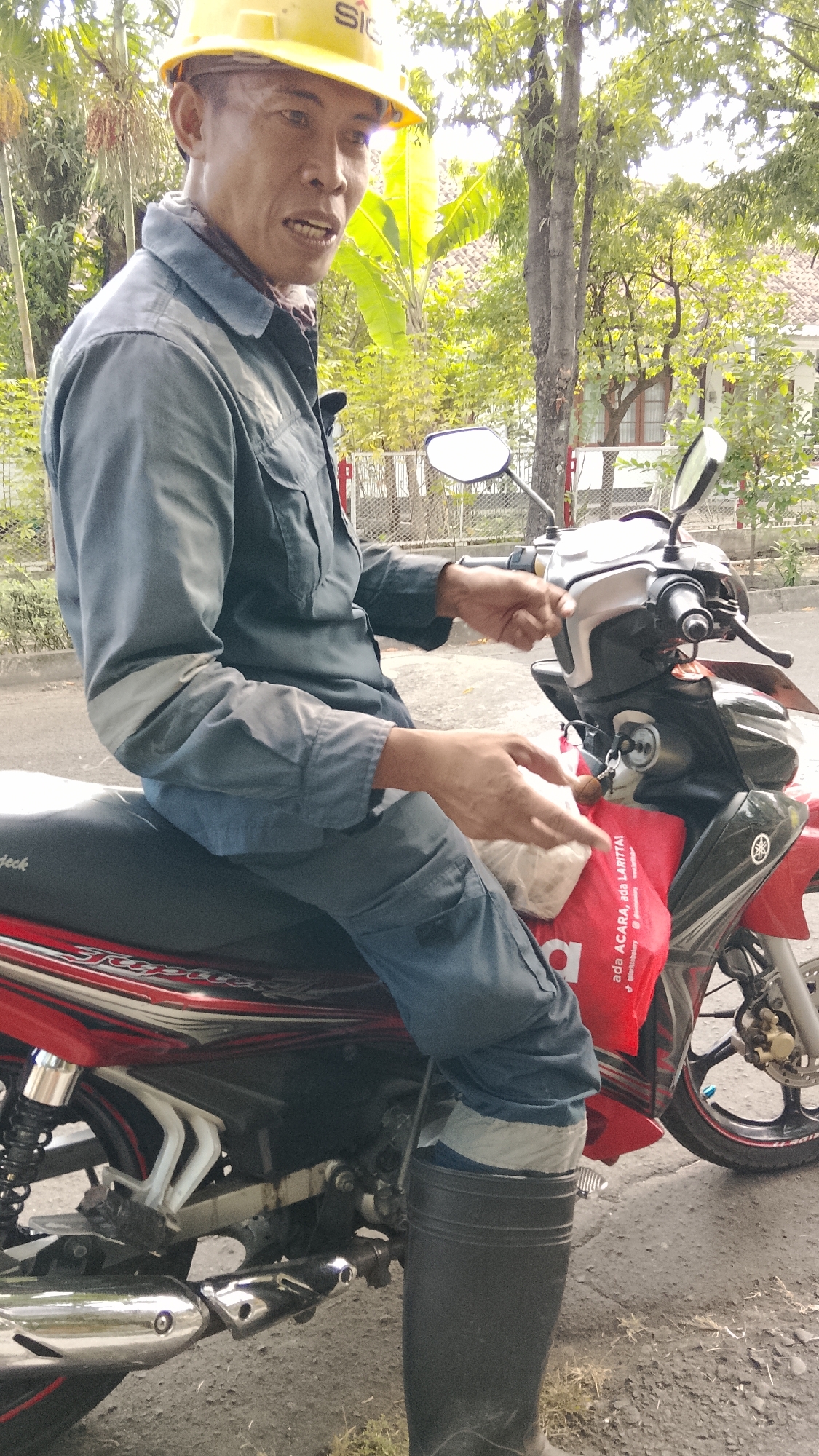 A middle-aged Asian man wearing a yellow hard hat and a blue work jumpsuit is sitting on a red and black motorcycle. The man has his mouth slightly open as if speaking, and he is looking towards the camera. He is holding a plastic bag filled with white items in his left hand, and his right hand is on the handlebar. The motorcycle is parked on an asphalt road, with a grassy area on the right side. In the background, there are trees and a building with a tiled roof, suggesting an outdoor setting in a residential or semi-rural area, potentially in Tlogobendung, Indonesia. The motorcycle has a visible exhaust pipe, a rear shock absorber, and a red bag hanging from the front.