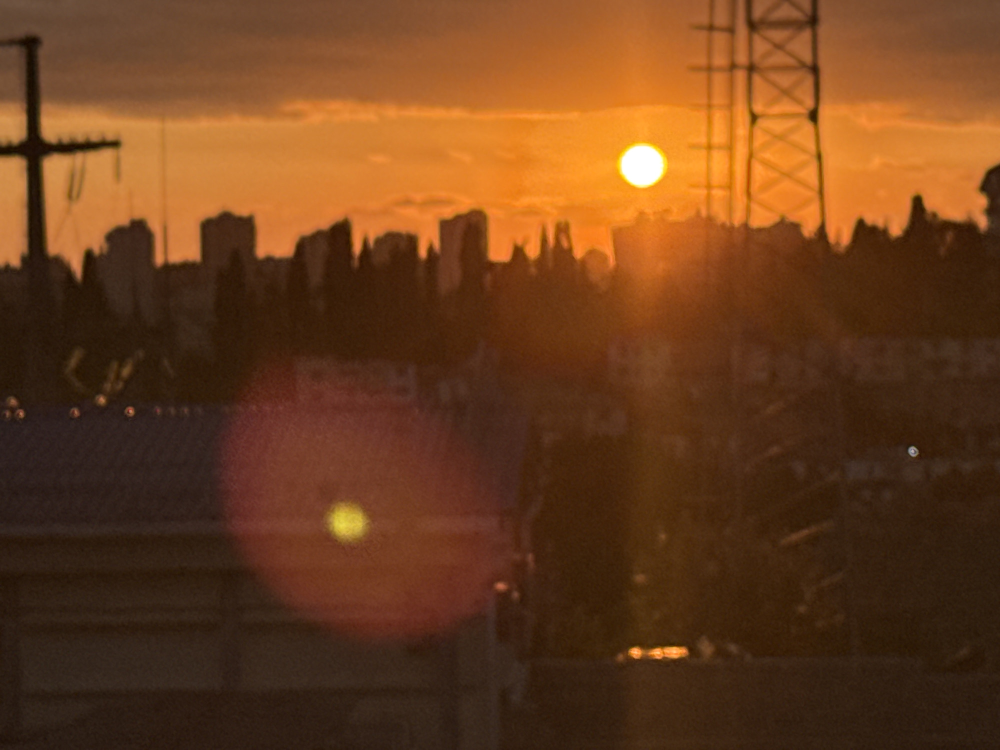 An extreme wide shot captures a sunset over a city, with a silhouette of buildings and a power line tower against the bright orange sky. The sun appears as a glowing orb, casting a warm light. In the foreground, a building's rooftop with visible horizontal lines is visible. A bright, circular lens flare with a yellow center is present in the lower left quadrant of the image. The overall scene is dark and hazy, suggesting atmospheric conditions or a low-quality image. The provided context suggests the location is Sochi, Russia.