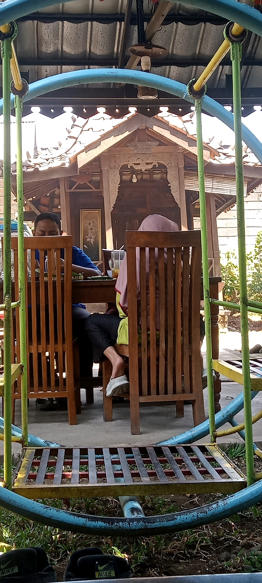 The image depicts a scene at a restaurant in Senggrong, Indonesia, where two individuals are seated at a wooden table. One person, wearing a blue shirt, is partially visible, appearing to be eating. The other individual, wearing a pink headscarf and a light-colored garment, is also seated and facing away from the camera. A glass of drink with a straw is placed on the table. Behind the table, a traditional wooden gazebo structure is visible, with intricate carvings and a tiled roof. The restaurant appears to be outdoors, with a grassy area and some vegetation in the background. In the foreground, part of a metal playground structure with blue and green tubing and a yellow frame is present, along with some black Nike sandals. The overall lighting suggests it is daytime, with sunlight filtering through the roof.