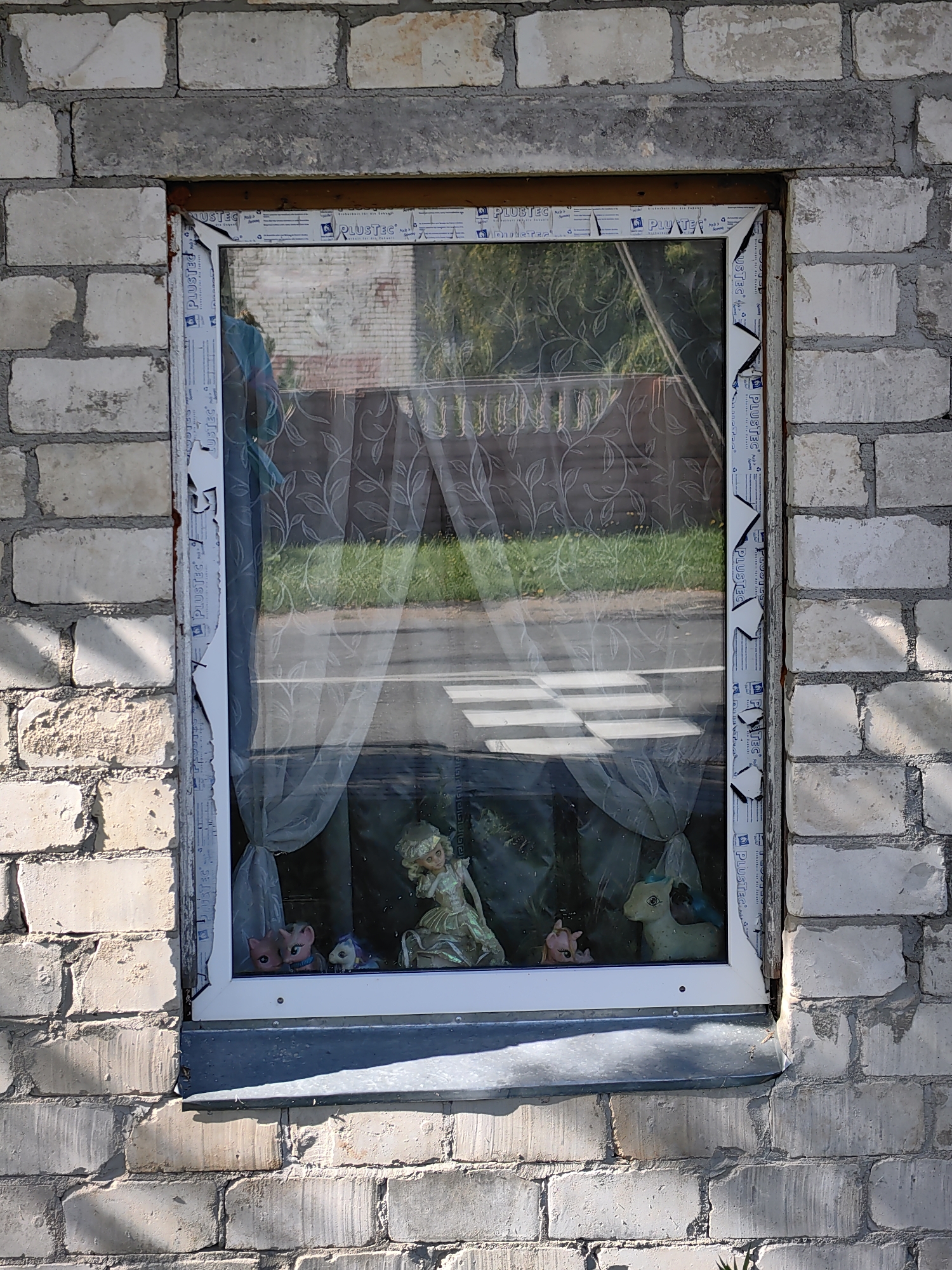 The image shows a close-up view of a window set into a light-colored brick wall. The window frame is white and appears to be made of vinyl, with blue and white stickers visible on its surface. The glass of the window reflects the outside environment, which includes trees and a patterned curtain hanging inside.
Inside the window, several figurines are visible. On the left, there are two pink figurines that resemble stylized ponies, with large eyes. To their right, there is a pale green figurine, also resembling a pony, with a turquoise mane and tail. In the center, a larger figurine of a blonde-haired doll with a flowing, iridescent dress stands prominently.
The brick wall surrounding the window is constructed from light-colored bricks, with visible mortar lines. The bricks are weathered, and some appear to be chipped or discolored. Shadows from the sunlight are cast across the wall and the windowsill, indicating it is daytime. The overall scene suggests a residential exterior in Myadzyel, Belarus.