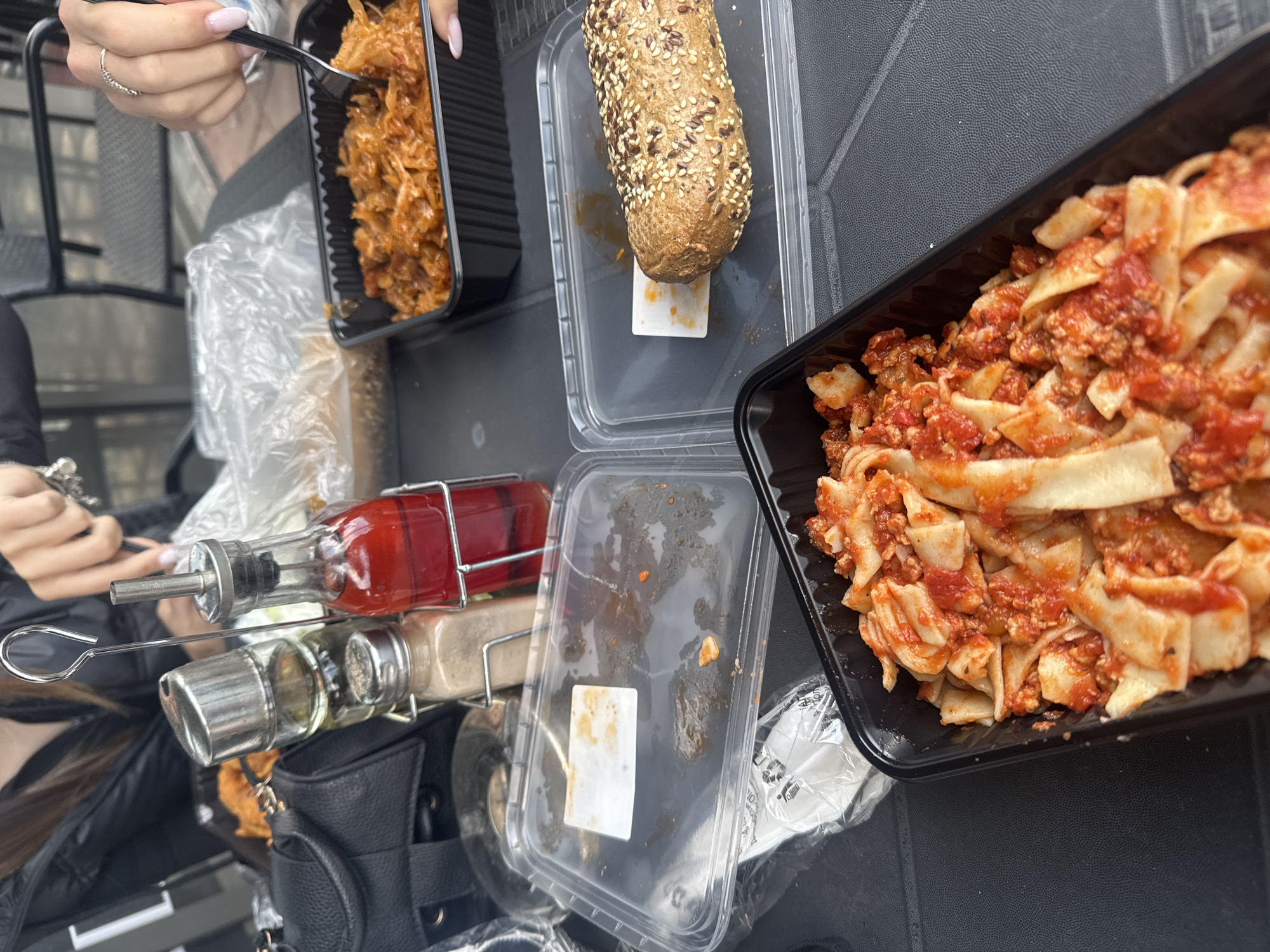 The scene depicts a meal in progress, presumably outdoors, likely in Plovdiv, Bulgaria. A person is using a black plastic fork to eat shredded meat from a black plastic container. Beside the container is a seeded roll in a clear plastic container. Next to the roll is another clear container that appears to be empty except for traces of food and a white label. To the right of the clear container is a bottle of red liquid with a metal dispenser and a two-tiered spice rack filled with glass jars. To the right of the condiment containers is a black plastic container filled with pasta and red sauce. The surface the containers are on is dark gray. The person eating the shredded meat is wearing a ring on their finger and is wearing light-colored clothing. The bag of the person is beside the containers.