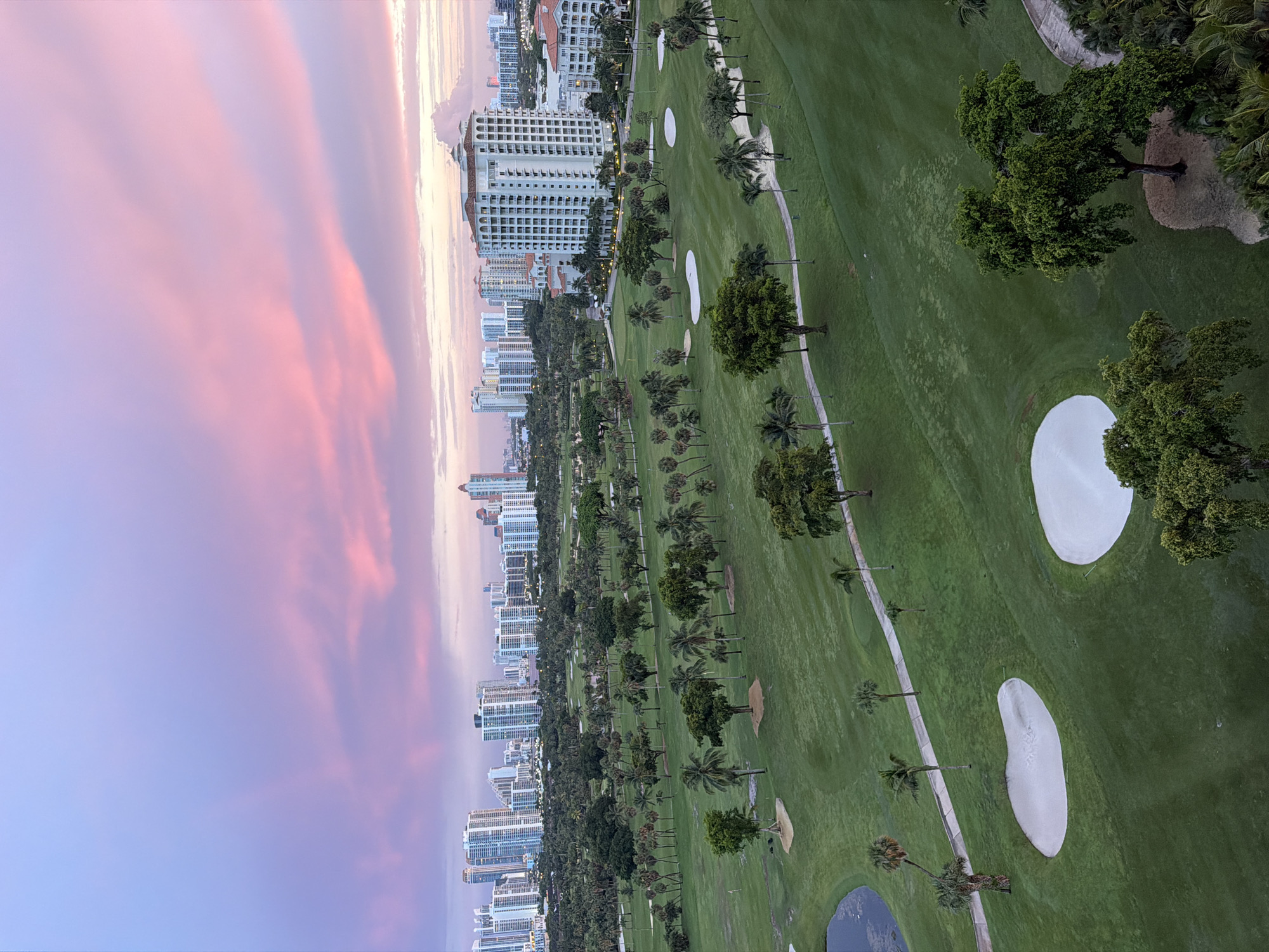 This is an aerial, high-angle, vertical shot of a golf course in Aventura, Florida, United States of America, during a sunset. The sky is a vibrant gradient of purple and pink, with wispy clouds. In the background, a dense cityscape of high-rise buildings, many appearing to be residential towers, stretches across the horizon. The golf course itself is lush green, with manicured fairways and strategically placed sand traps. Several palm trees and other types of trees are scattered across the landscape, adding depth and texture. A paved path winds through the course. There are no people visible in the image.
