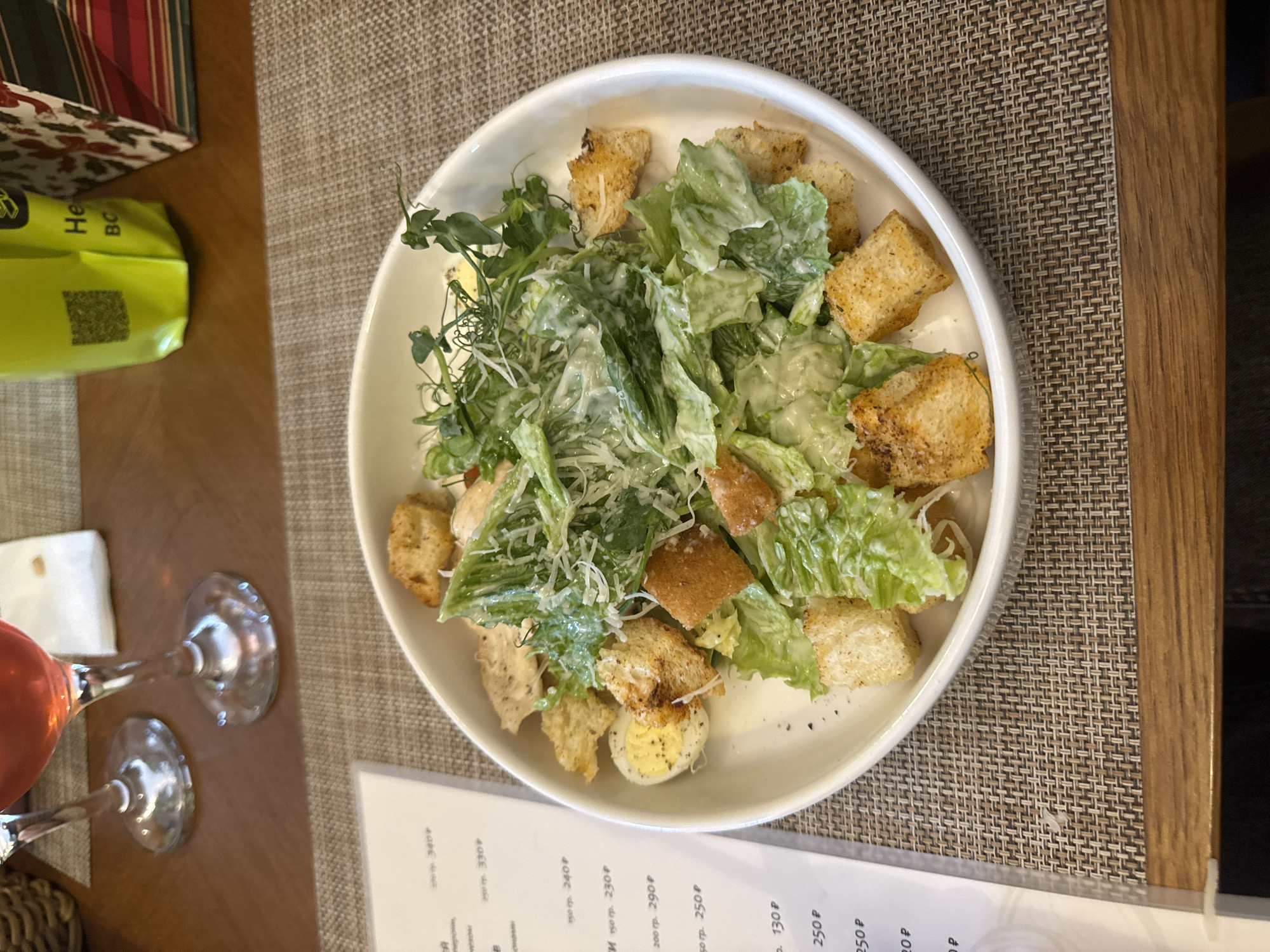 The image depicts an overhead view of a dining table in a restaurant located in Perm, Russia. A white, circular bowl centrally features a Caesar salad composed of romaine lettuce, numerous cuboid toasted croutons, finely grated white cheese, a creamy white dressing, multiple pieces of cooked chicken, and one half of a hard-boiled egg seasoned with black pepper. The salad is garnished with a cluster of green microgreens. The bowl rests on a rectangular woven placemat exhibiting a brown and beige basketweave pattern. The table surface is polished wood. To the left of the bowl, a laminated or protected menu is visible, displaying Cyrillic text and prices in Russian Rubles (e.g., "340 Р", "330 Р"). In the upper left background, two stemmed glasses, one containing a reddish-pink liquid, a folded white napkin, and a segment of a wicker basket are present. In the upper right background, a bright neon green flexible packaging item, possibly a bag, displays a partial QR code and Cyrillic characters. Adjacent to this is a decorative rectangular box or bag with red, white, and green patterns. No individuals are visible in the frame.