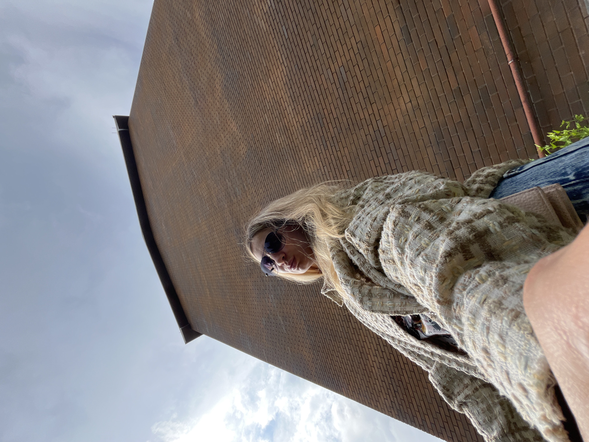 A low-angle outdoor shot shows a blonde woman wearing sunglasses and a textured, neutral-colored jacket standing in front of a large, tall brick building under a cloudy sky. She is looking away from the camera with her lips pursed. The building, which appears to be in Pionersky, Russia, dominates the background, stretching upwards into the sky. The perspective emphasizes the scale of the building.