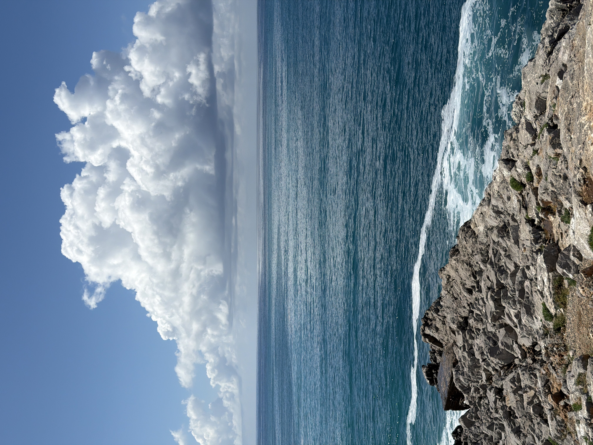 The image captures a vibrant coastal landscape in Bečići, Montenegro, under bright daylight. The scene is dominated by three main elements: a vast blue sky with prominent clouds, an expansive ocean, and a rugged rocky cliff.
The upper portion of the frame features a clear blue sky. On the left side, a large, impressive formation of fluffy white cumulus clouds extends, showcasing intricate details, shadows, and brilliant highlights from the sun. The clouds appear voluminous and well-defined against the azure backdrop.
Below the sky, the deep blue-green waters of the sea stretch out to the horizon. The ocean's surface is textured with countless ripples and small waves, reflecting the sunlight and creating a shimmering effect. The water appears calm in the distance but more dynamic closer to the shore.
On the right side of the image, a dramatic, weathered rocky cliff face descends steeply into the sea. The grey rock formations are rough and jagged, with sparse patches of green vegetation clinging to the ledges and crevices. Along the base of the cliff, white foamy waves crash and dissipate into the ocean, indicating active sea movement.
The clear visibility and strong illumination suggest it is mid-day with sunny weather. There are no people, man-made objects, or visible text in the scene. The overall impression is one of natural grandeur and tranquility.