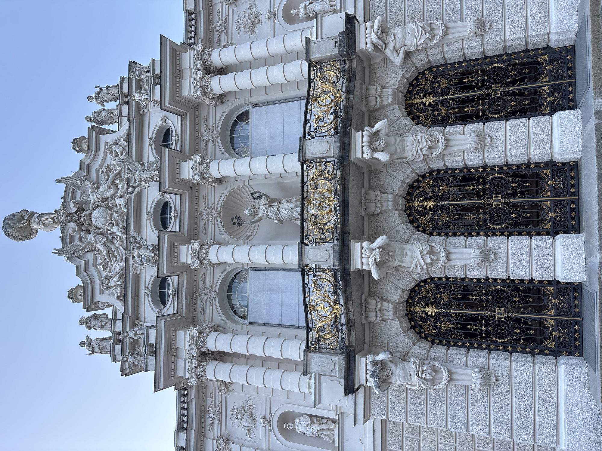 The image shows the exterior of a grand white building with ornate architectural details, likely a palace or a historic monument, situated in Ettal, Germany. The building features a detailed facade with multiple levels, characterized by classical columns, sculptures, and decorative friezes. A prominent balcony with intricate gold railing is visible in the center, supported by muscular statues. Below the balcony are large, arched doorways with decorative black and gold metal gates. The sky above is a clear, pale blue, suggesting it is daytime, possibly early morning or late afternoon due to the soft light. The overall impression is one of opulence and historical significance.