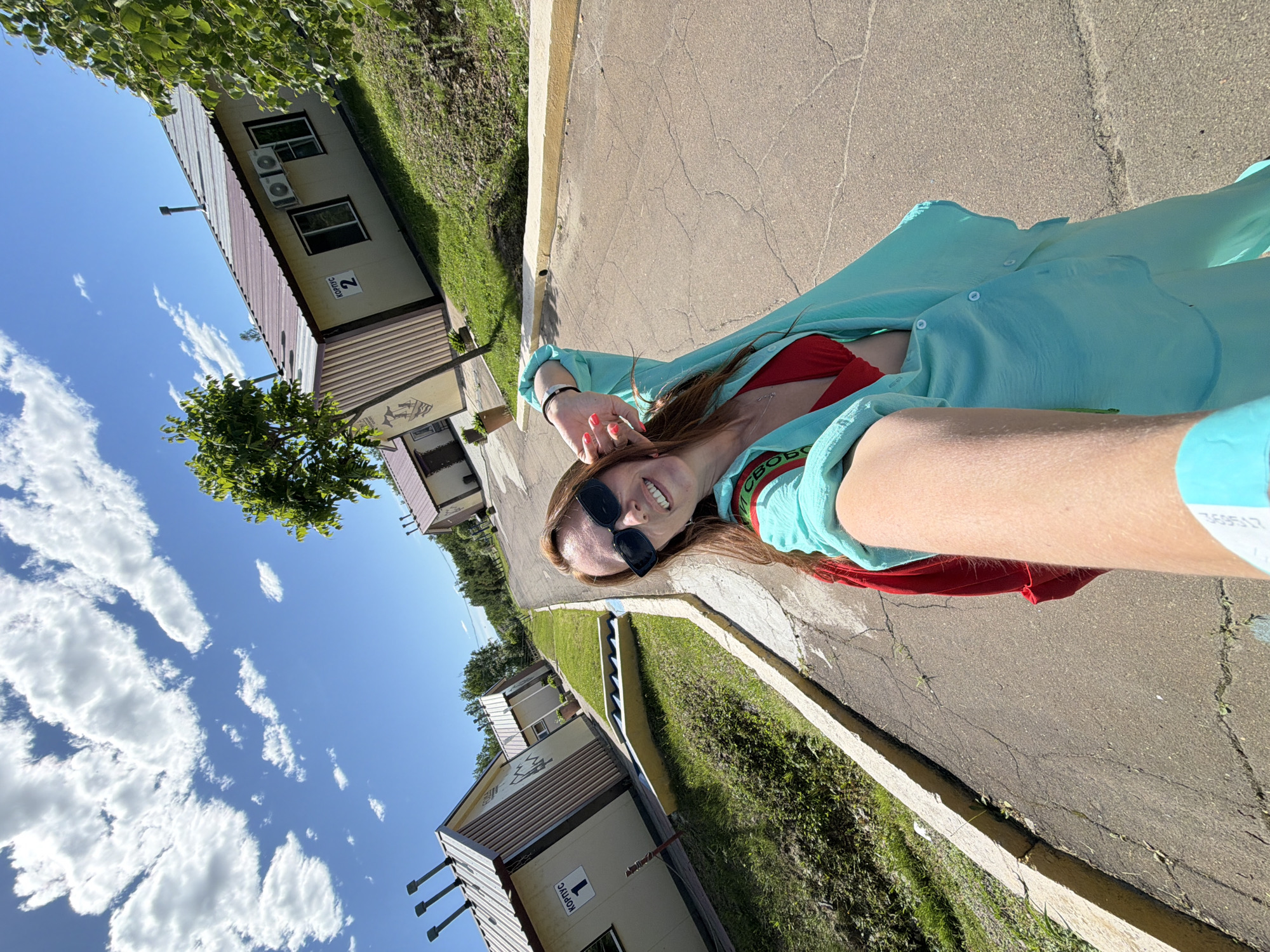 A woman with long brown hair and sunglasses is taking a selfie in an outdoor setting. She is wearing a red bikini top and a light blue open shirt, with a red backpack visible. The background shows a row of small beige buildings with brown roofs, labeled as "корпус" (building) numbers 1 and 2. These buildings appear to be part of a resort or camp. A paved road runs alongside the buildings, with grassy areas and trees. The sky is blue with scattered white clouds. The scene suggests a warm, sunny day in a recreational area. The image was taken in the city of Birobidzhan, Russia.