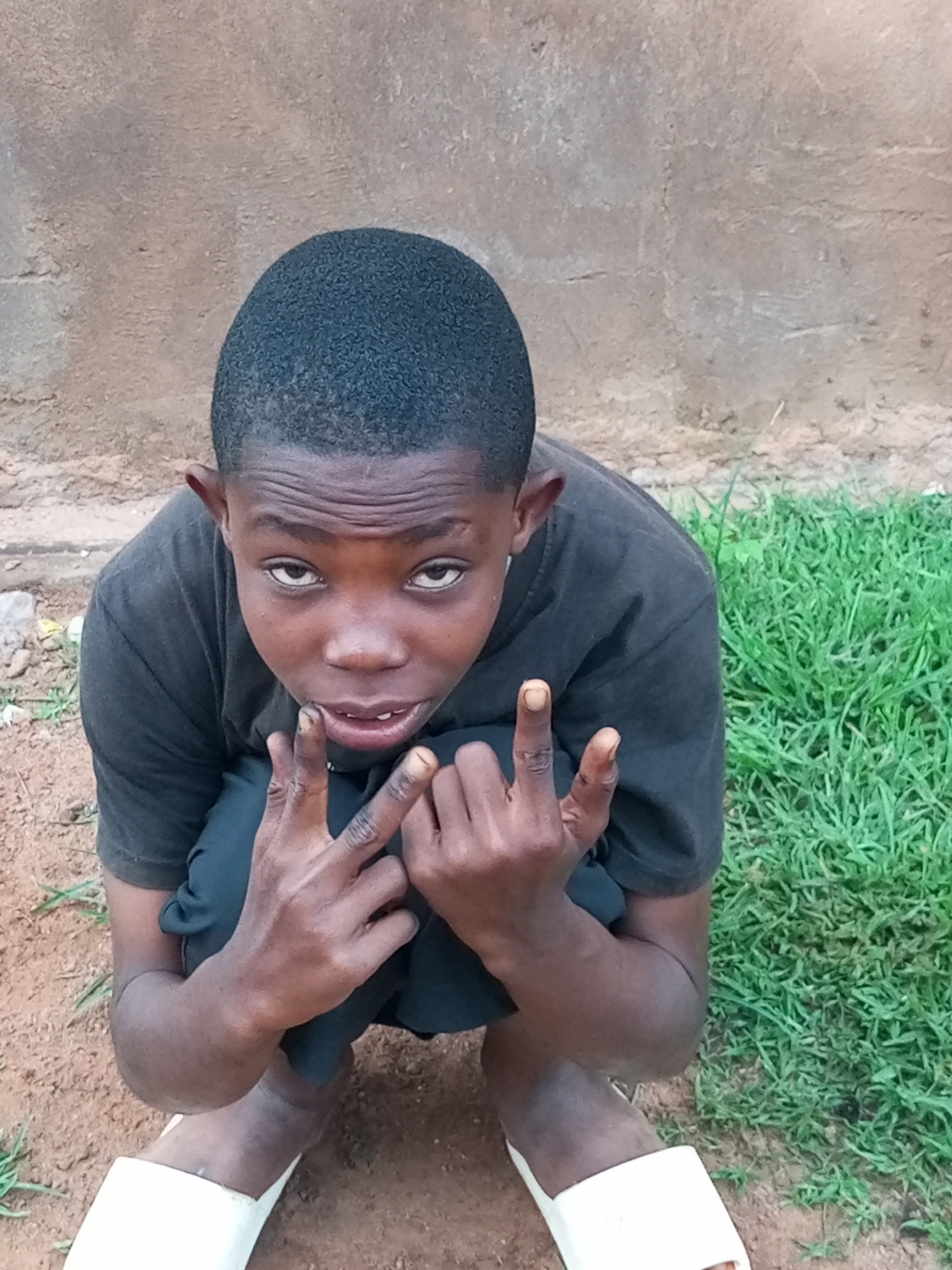 A young Black boy is captured in a medium, high-angle shot, crouching down outdoors. He is wearing a plain black t-shirt and black shorts. His hands are raised in front of him, with his index fingers and thumbs extended, forming a "gun" gesture. The boy has short, dark hair and is looking directly at the camera with a neutral expression. The background consists of a rough, textured wall and a patch of green grass. The ground is composed of dirt and a few scattered stones. The scene appears to be located in a casual outdoor setting, possibly a courtyard or alleyway in the city of Tempure, Nigeria.
