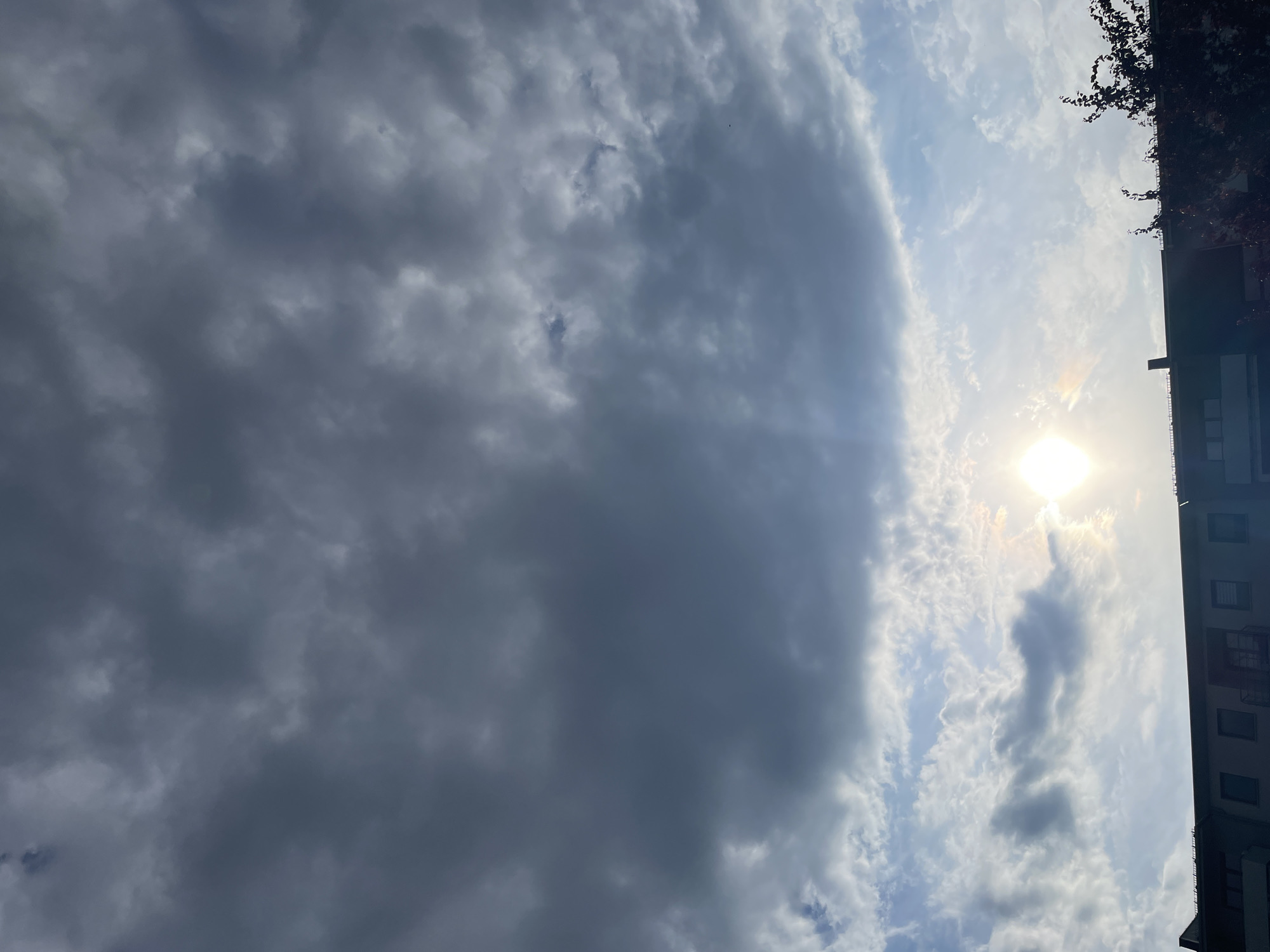 The image shows a daytime sky partially obscured by clouds. A section of a building is visible on the right side of the frame, with several windows. Branches from a tree or bush extend from the top right corner. The sun is visible through a break in the clouds, creating a bright spot in the sky. The majority of the sky is covered with darker, denser clouds. The location of the image is Unknown, Unknown.