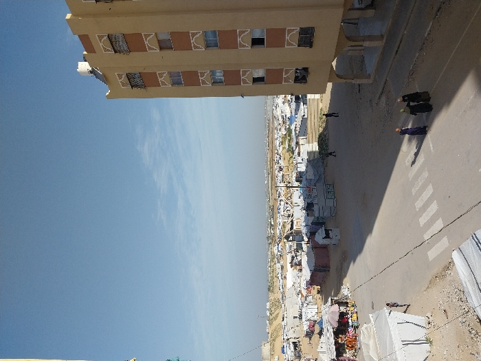 A high-angle shot shows a city street adjacent to a tall building. Three women walk across a zebra crossing on the street. Behind them, a line of tents forms a camp-like area. In the background, more buildings stretch into the distance under a clear blue sky. The overall scene suggests a densely populated urban area with a mix of permanent and temporary structures.