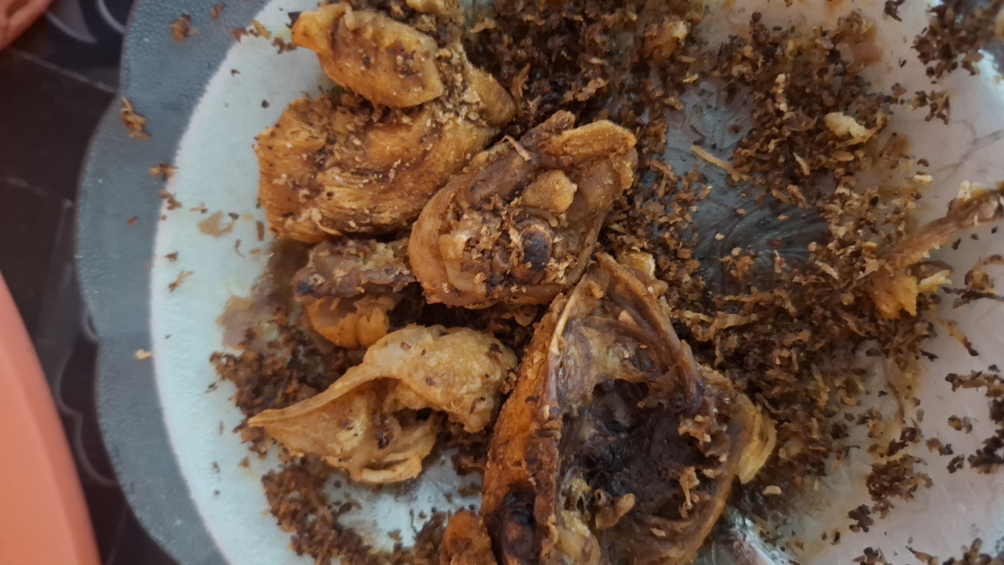 A close-up, overhead shot shows several pieces of fried food on a speckled, light-blue plate. The food appears to be mostly fried chicken, with some pieces showing skin and bone. The fried pieces are golden brown and covered in a light dusting of crumbs or seasoning. The plate is set on a dark, patterned surface, possibly a table. In the background, a blurry, curved, orange object is visible, suggesting it could be part of another dish or a container. The overall scene is dimly lit, with the focus entirely on the food. This scene is located in Ciharashas, Indonesia.