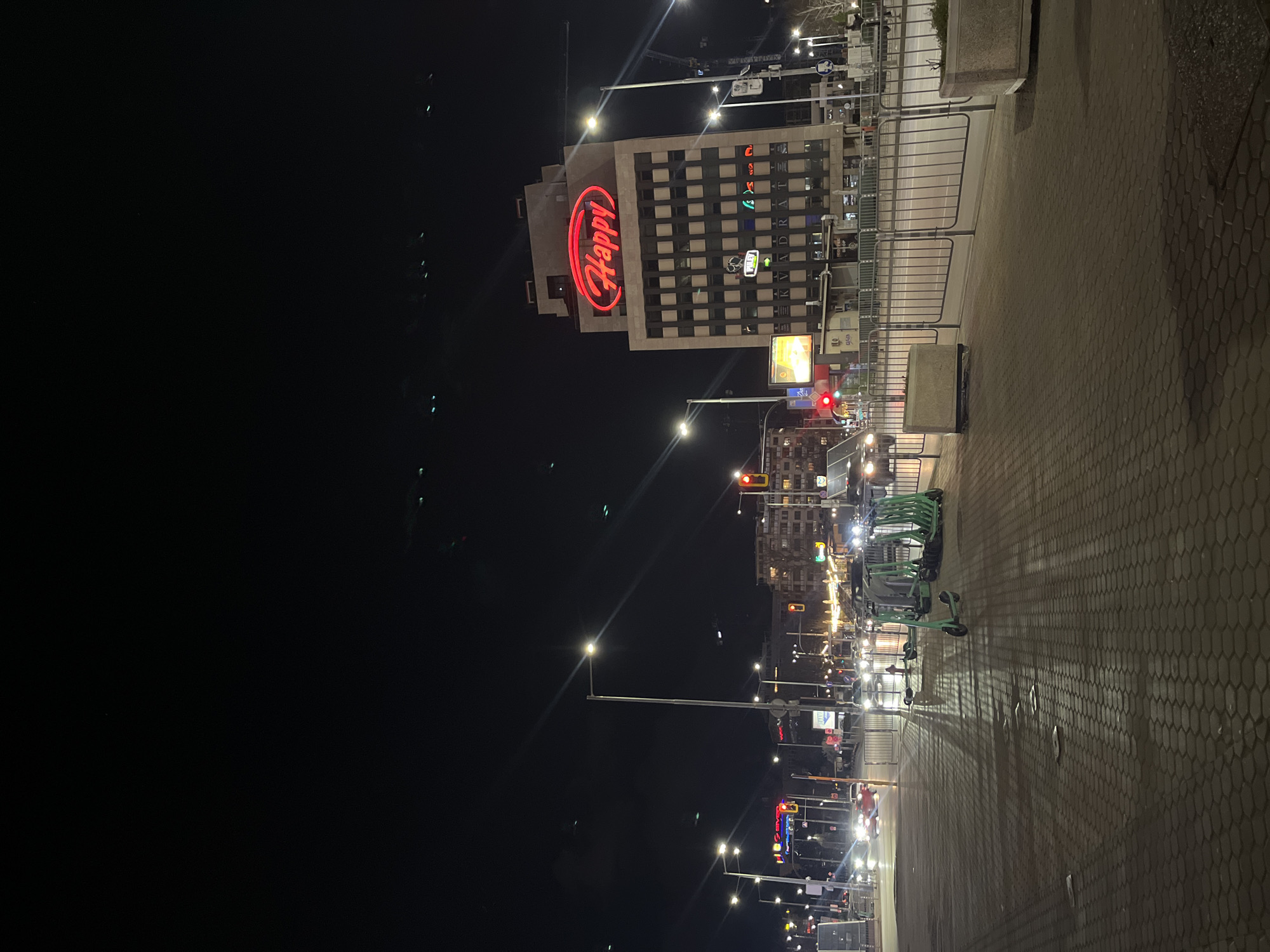 The image depicts a nighttime street scene in Sofia, Bulgaria. Dominating the view is a building with a large, illuminated "Happy" sign in red. Streetlights illuminate the road and sidewalk, where several green scooters are parked. Cars and pedestrian traffic are visible further down the street. The sky is dark, and the overall impression is of an urban setting at night.