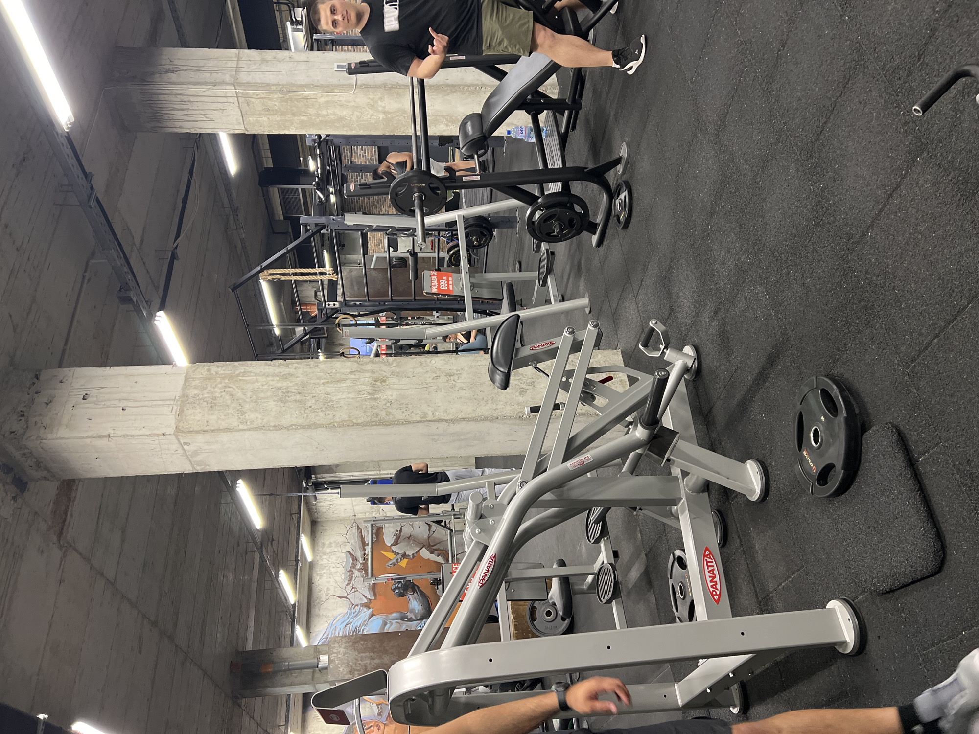 The image shows the interior of a gym. There are several pieces of exercise equipment, including weight benches, plate-loaded machines, and barbells. People are visible, some appearing to be exercising. The gym has a concrete and industrial aesthetic, with visible structural elements and bright overhead lighting. The presence of a "PROMO PRICE" sign written in Bulgarian language suggests that the location is within Bulgaria, likely Sofia. The specific location cannot be determined from the image alone.