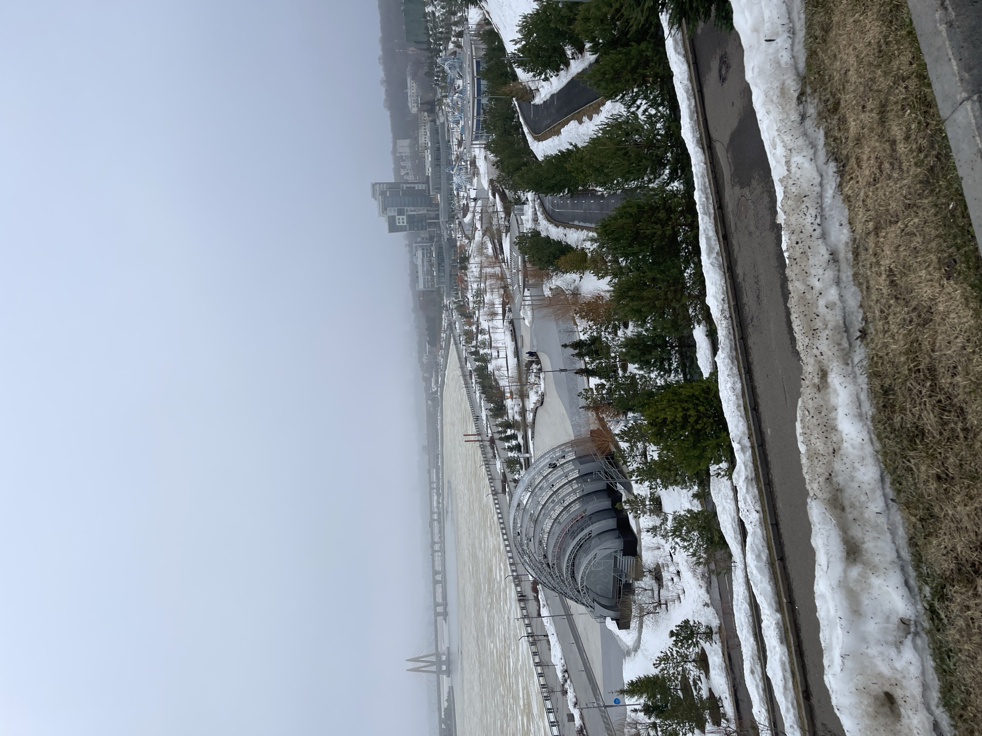 The image shows an elevated perspective of Kazan, Russia, with a heavy fog obscuring the distant cityscape. The foreground features a riverbank with snow patches, bare trees, and evergreen trees. A distinctive, large metal structure with a semi-circular, layered design dominates the midground. Adjacent to this structure is a paved pathway. In the background, the river appears wide and muddy with two cable-stayed bridges visible through the fog. The photo is taken in winter, judging by the snow cover along the side of the road. The scene appears deserted; no people are visible.