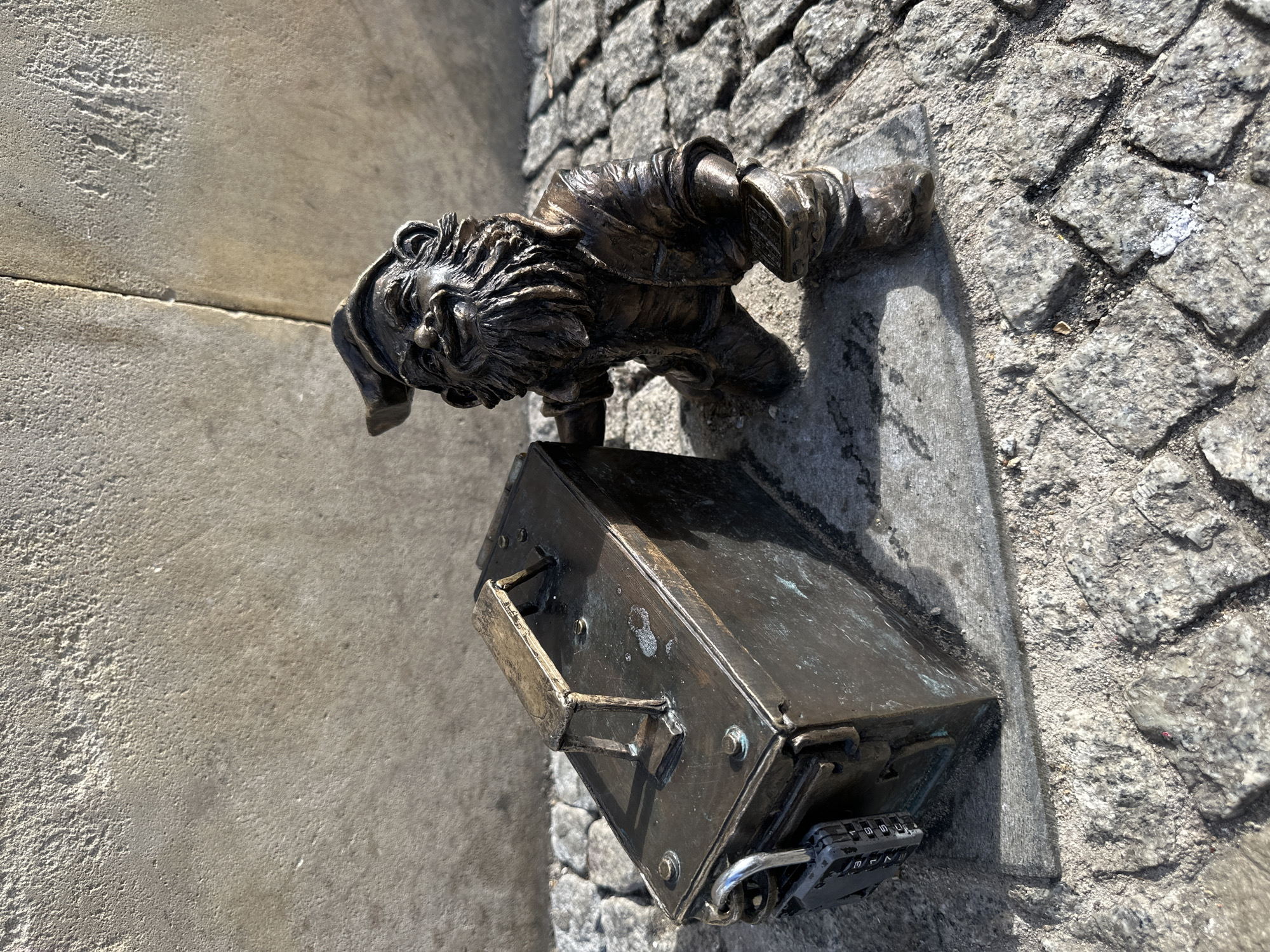 A bronze dwarf statue with a beard and cap is leaning over a bronze toolbox on a cobblestone street. The toolbox is secured with a combination padlock. The statue and toolbox are located in Wrocław, Poland, as these statues are common throughout the city. The backdrop consists of cobblestone and a concrete surface, likely a curb or building foundation.