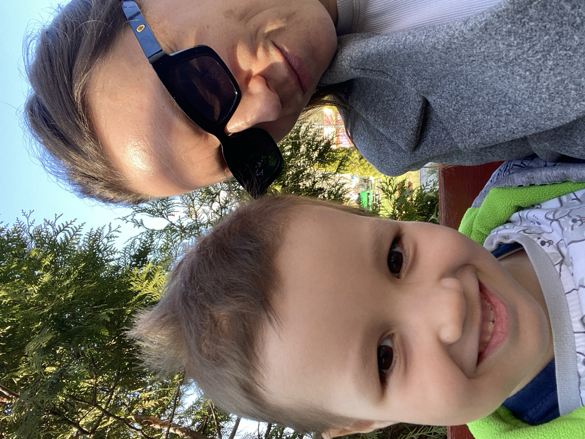 A low-angle selfie-style photo features a woman and a young boy. The woman is positioned at the top of the frame, wearing dark sunglasses and a grey sweater. Below her is a smiling young boy. The background shows outdoor greenery, possibly trees, and hints of a structure, indicating a park or outdoor recreational area. The location is Chaikovsky, Russia.