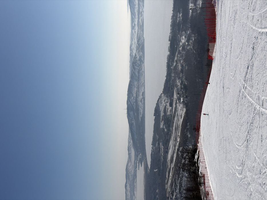 An elevated view captures a winter landscape from a ski slope overlooking a large, frozen body of water identified as being in Zelenaya Polyana, Russia. The foreground features a snow-covered ski run with visible ski tracks. To the left, a portion of a dark, rocky outcrop with sparse trees is visible. Bright orange safety netting or fencing delineates the edges of the ski slope on both the left and right sides. Two individuals are present on the slope: one in red attire stands mid-slope on the left, facing downhill, while another figure in red is further down and to the right, also on the slope.
Beyond the immediate slope, the terrain slopes downwards, transitioning into a snow-dusted, wooded area with visible buildings and infrastructure, suggesting a settlement or resort facilities. A vast, flat, ice-covered lake extends across the mid-ground, reflecting the clear, pale blue sky above. Distant, snow-capped hills and mountains frame the background, with some exhibiting visible structures such as communication towers on their peaks. The sky is clear and bright, indicating a sunny day. The overall scene depicts an active winter sports area with a panoramic view.