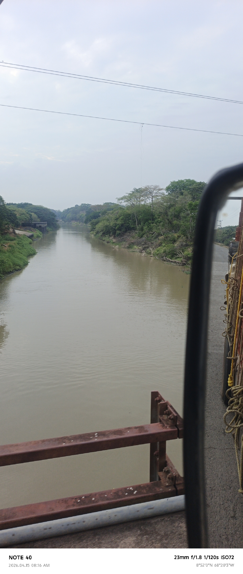 This eye-level photograph presents a river scene in what is likely El Baúl, Venezuela, as indicated by the GPS coordinates (8°52'0"N 68°20'3"W) embedded in the image's metadata. The view is partially obstructed by elements of what appears to be a vehicle or bridge structure, including rust-colored metal beams in the foreground and a blurred side mirror and tied ropes visible on the right side.
The river, occupying the central portion of the frame, has a muddy brown color. Both banks are lined with green vegetation, including trees and shrubs, contributing to a lush, natural setting. A concrete structure or building is seen on the left bank in the distance. Overhead, power lines stretch across the sky. The sky itself is overcast. The image's metadata specifies that it was taken with a 23mm lens at f/1.8, with an exposure of 1/120s, and ISO72.