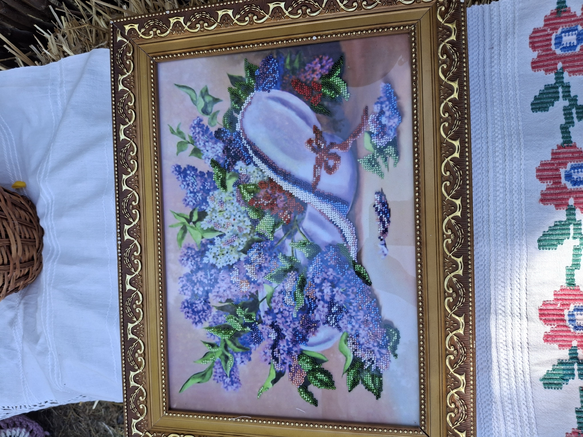A framed beaded picture of a floral still life featuring a bouquet of lilacs spilling from a light purple hat with a red ribbon. The picture is resting on a white embroidered cloth, which is on top of a surface covered in straw and dried grass. In the upper left corner of the image, a portion of a woven basket is visible. The scene is outdoors, likely in a rural setting in Roghi, Moldova.