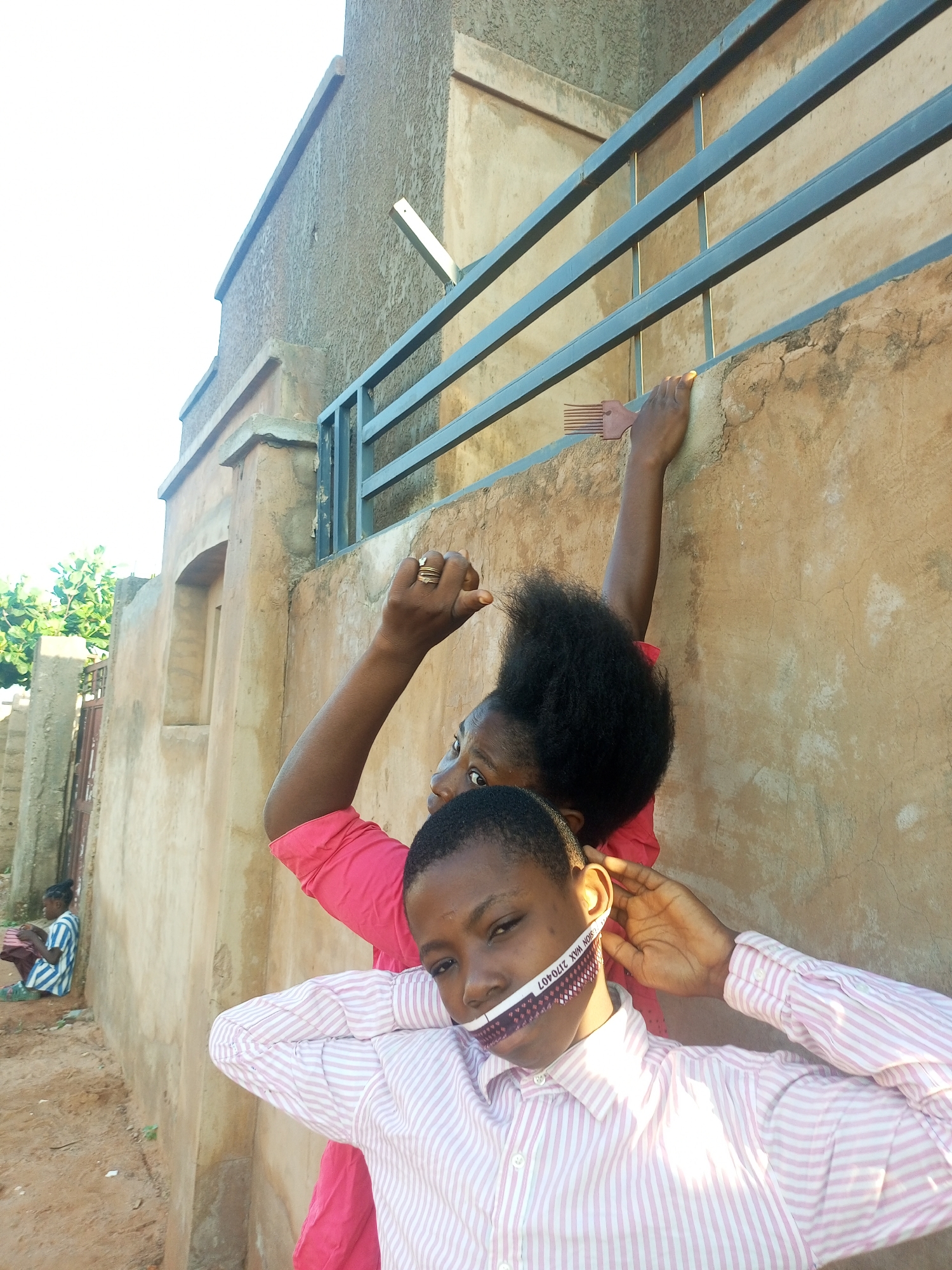 In Tempure, Nigeria, a person in a striped pink and white collared shirt is positioned in the foreground, looking directly at the camera with their mouth covered by a strip of fabric displaying a zigzag pattern and text that reads "VISION WAX 270407". Their left hand is raised, holding the fabric in place near their ear, while their right arm is bent at the elbow. Behind them, another person wearing a bright pink top is reaching upwards, their left hand touching a metal railing on a textured, light brown wall. This second person is looking towards the left of the frame, and their right hand is visible holding a comb with red prongs. In the background, to the left, another individual is seated on the ground, facing away from the camera and looking to the left. This seated person is wearing a horizontally striped blue and white outfit and has a cloth folded in their lap. The environment appears to be outdoors, with sandy ground and vegetation visible to the far left. The wall has a rough, stucco-like texture.