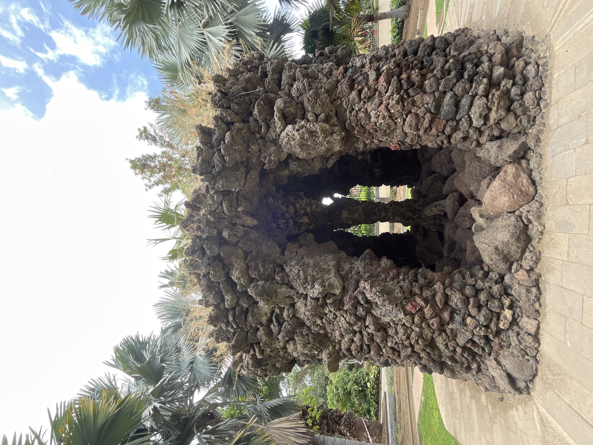 Here's a detailed description of the image:
**Location:** This appears to be a structure at Parque Doramas in Las Palmas de Gran Canaria, Spain.
**Subject:** The focal point is a man-made structure constructed of volcanic rock. The structure has a roughly rectangular shape with openings on both sides, creating a tunnel-like effect.
**Objects:** The structure is primarily composed of dark, porous volcanic rocks, varying in size. Some vegetation is growing from the top of the structure. The surrounding environment includes palm trees.
**Environment:** The scene is outdoors with a bright, daytime setting. The sky is partially visible with clouds. The ground consists of pavement and grass.