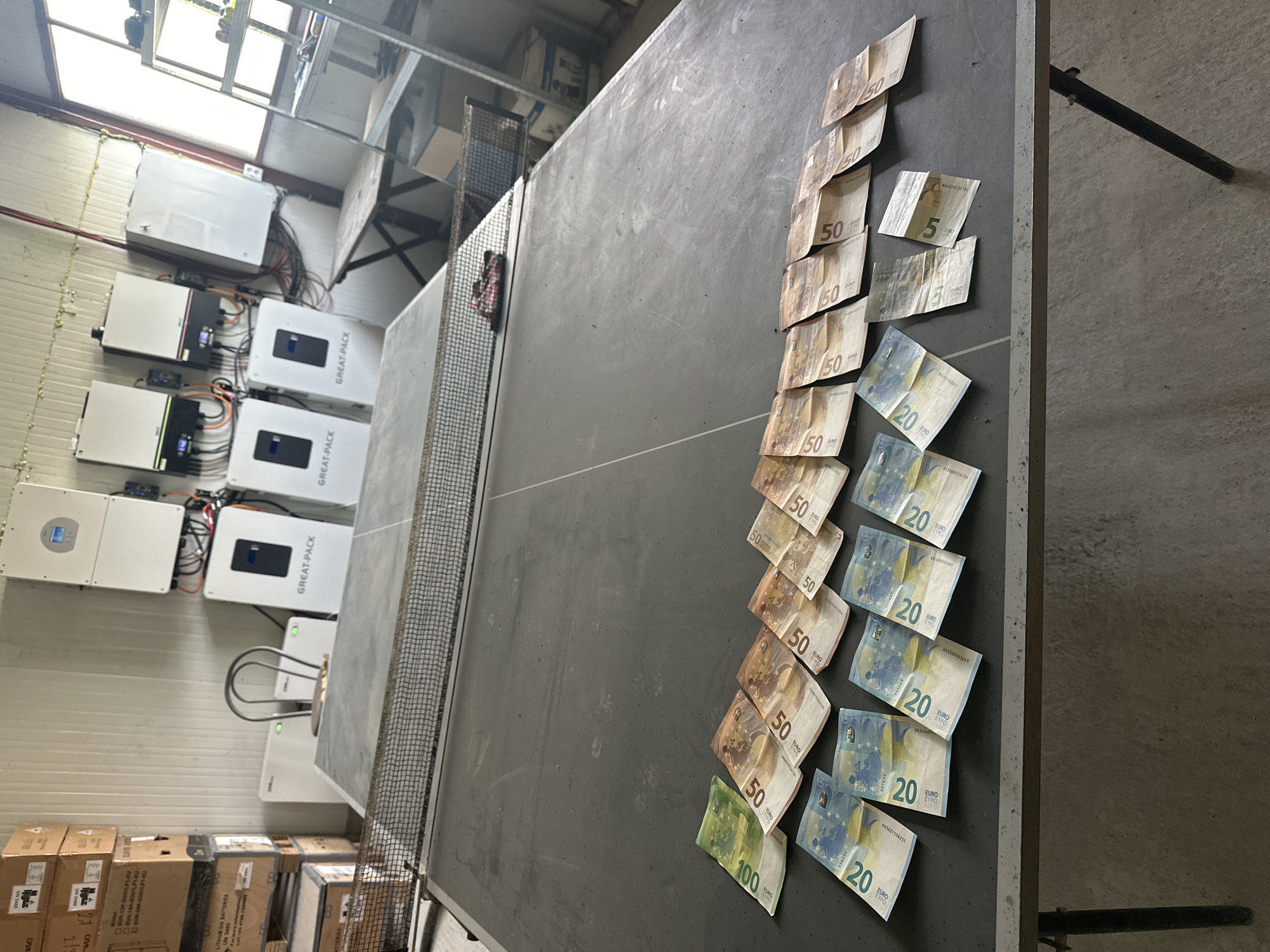 This media file captures an indoor scene, likely a warehouse or industrial space, located in Saraia, Bulgaria. The image, illuminated by natural light from overhead windows, features a dark grey ping-pong table prominently in the foreground and midground.
Spread out neatly on one side of the ping-pong table are various Euro banknotes. These include one 100 Euro note, twelve 50 Euro notes, seven 20 Euro notes, and two 5 Euro notes, totaling 850 Euros.
In the background, mounted on a light-colored wall, are multiple white, rectangular electronic units, likely solar inverters or battery storage systems, with visible cabling connecting them. Four of these units are clearly branded "GREAT-PACK," and one bears the model number "SUN-12K-0001LP14U." Below these units, stacks of cardboard boxes are visible, several of which are labeled "LITHIUM ION BATTERIES" and "UN 3460." Additional text on boxes includes "US," "DO," "DAV," and "21."
The setup, with energy equipment and battery storage boxes alongside a display of cash, suggests a commercial or operational environment related to renewable energy or power solutions. No individuals are visible in the image.