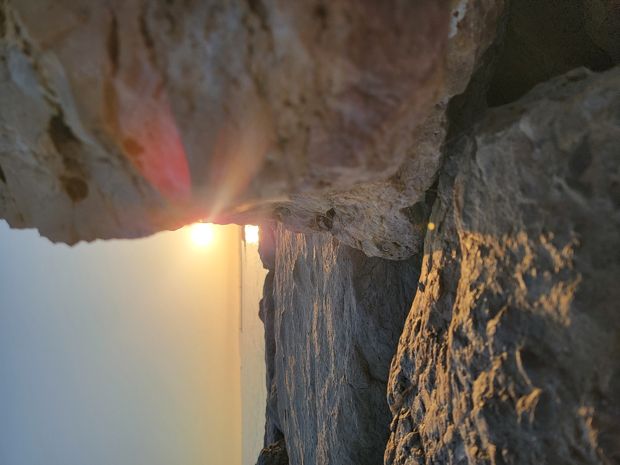 A low-angle, close-up shot of rocks on a shoreline is captured during sunset in Quarteira, Portugal. The sun, a bright orb near the horizon, casts a warm glow across the scene, creating lens flares that streak across a large, light-colored rock in the foreground. The rock, which dominates the right side of the frame, is textured and rough, with some darker mineral deposits. In the middle ground, the gentle waves of the Atlantic Ocean meet a rocky shore, with faint structures visible in the distance on the horizon. The foreground rocks are rough and grey, with warm light illuminating their surfaces from the setting sun. The sky transitions from a clear blue at the top to a soft orange and yellow near the horizon. There are no people or animals visible in the image.