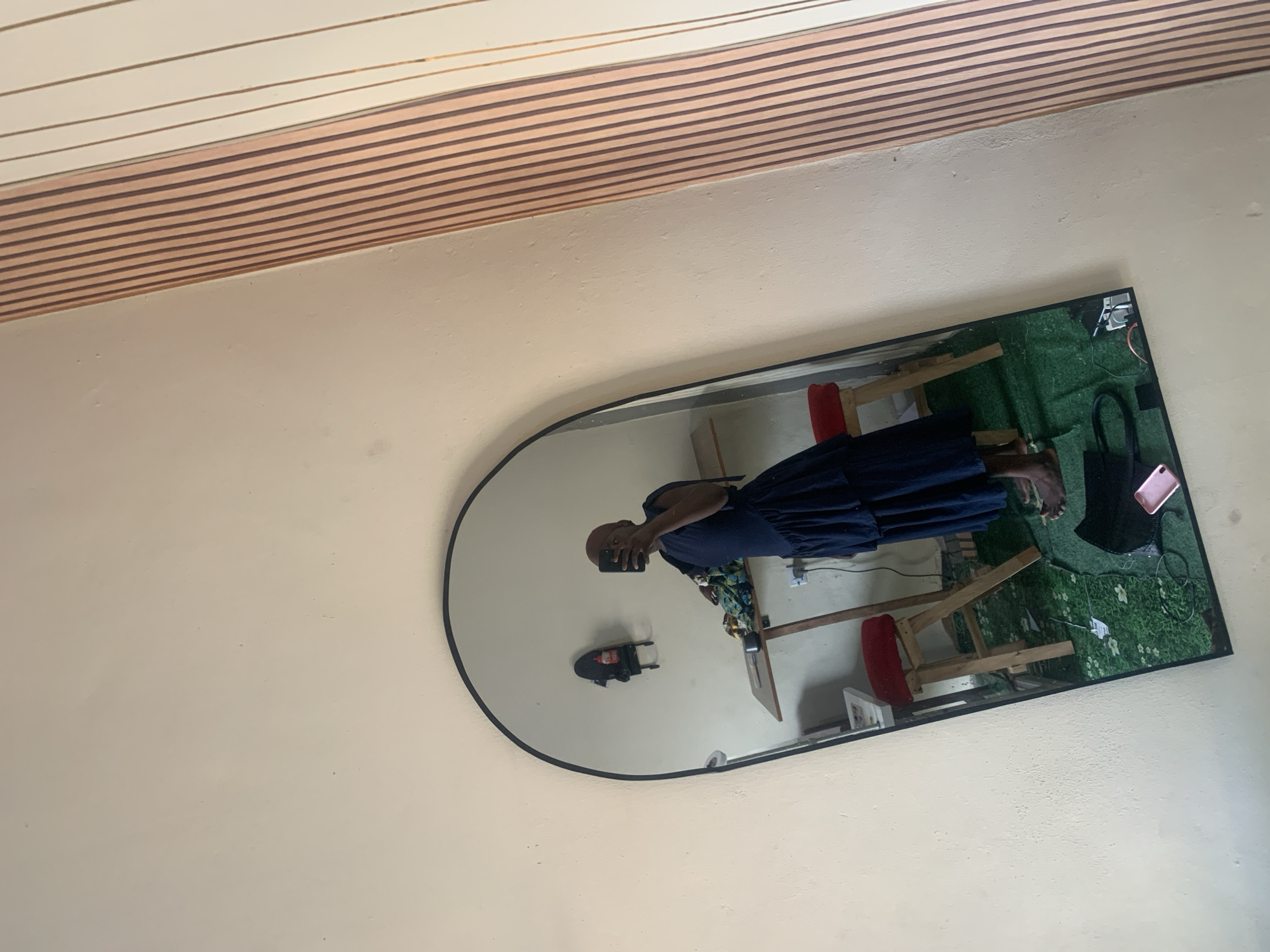 A person is taking a mirror selfie in a room with a large, arched mirror. The person is wearing a dark blue ruffled dress and is holding a black smartphone to capture the reflection. In the mirror's reflection, a wooden table with items on it, two red stools, and a faux grass rug are visible on the floor. A shelf with a bottle and other items is mounted on the wall behind the table. The room appears to be a domestic space, possibly in the city of Jauro Yinu, Nigeria, given the context. The wall to the right of the mirror has a striped wallpaper in shades of cream and light brown.